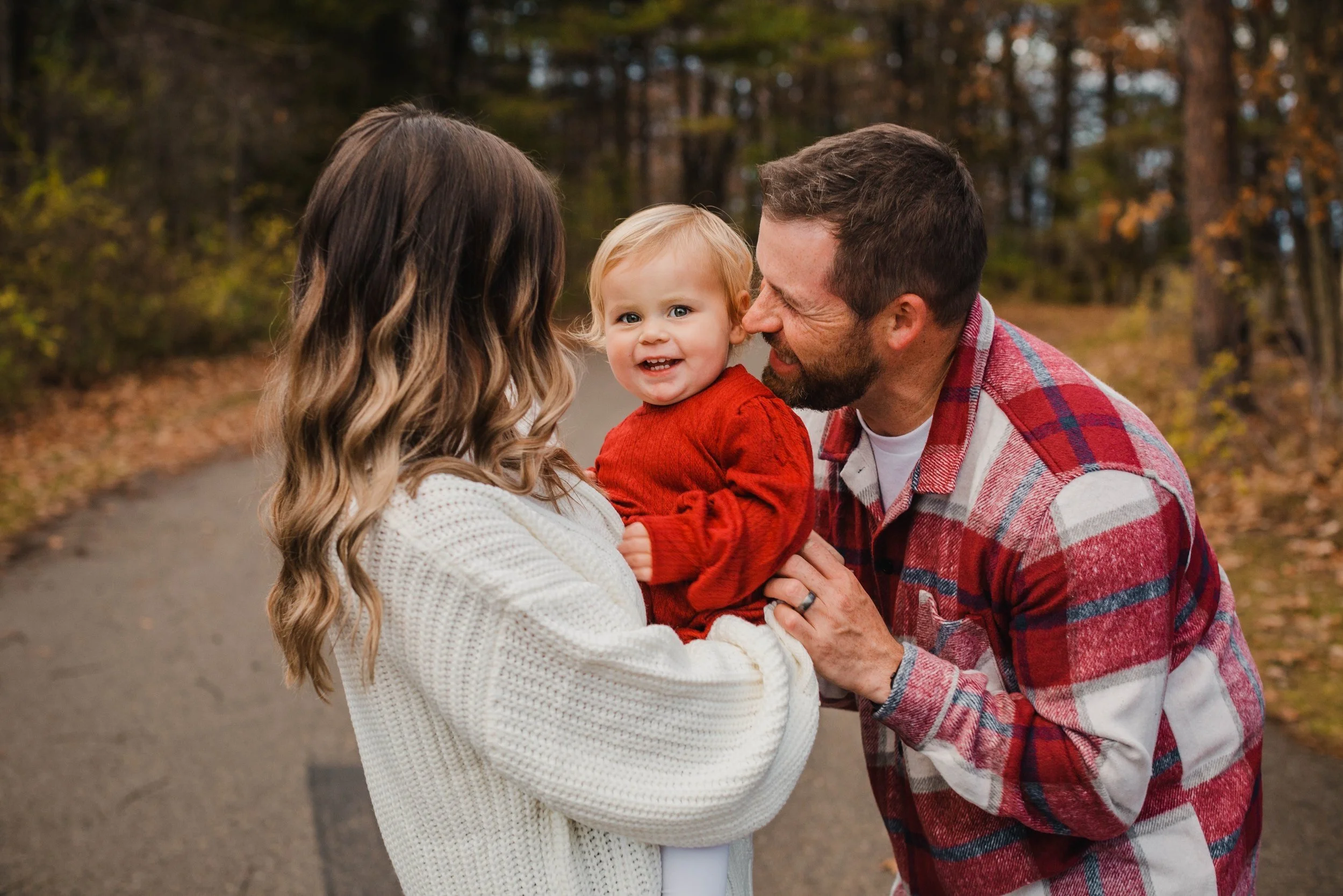 A family of three outdoors during fall, with the mother holding a baby girl in a red sweater, while the father, wearing a red plaid shirt, smiles and interacts with the baby.