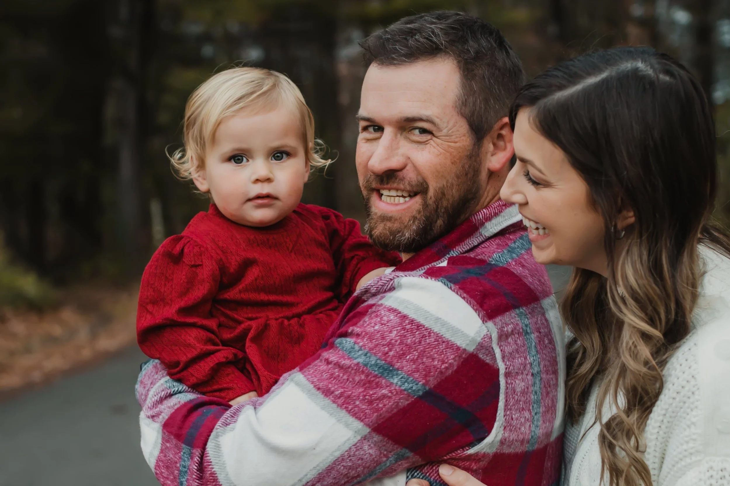 A family outdoors with a man holding a young girl in a red dress, while a woman smiles beside them on a wooded path.