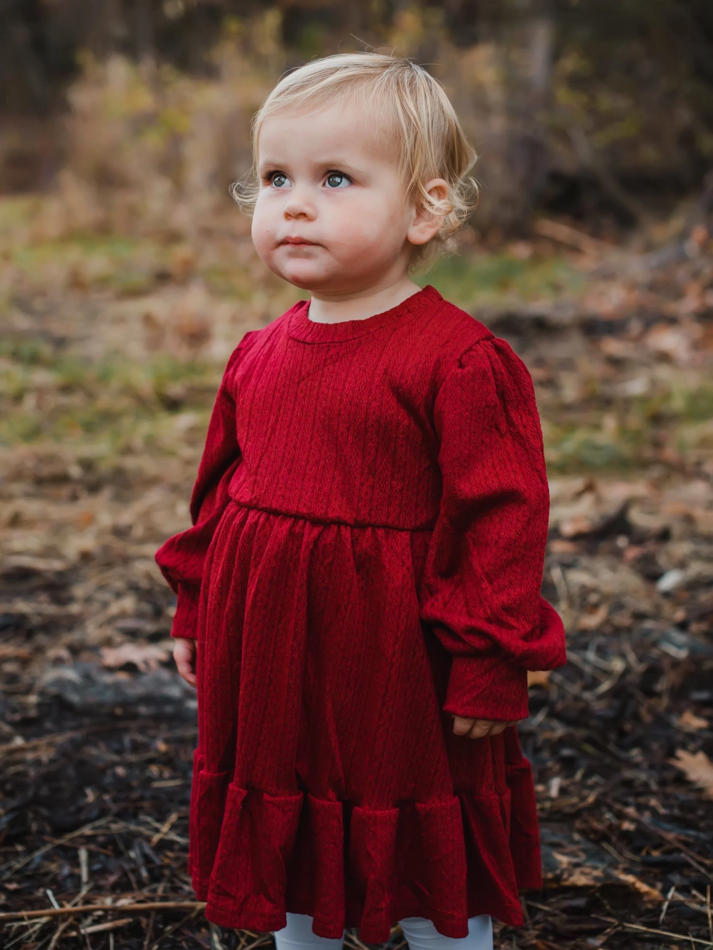 A young girl with blonde hair, blue eyes, wearing a long red dress, standing outdoors on a fall day with fallen leaves and trees in the background.