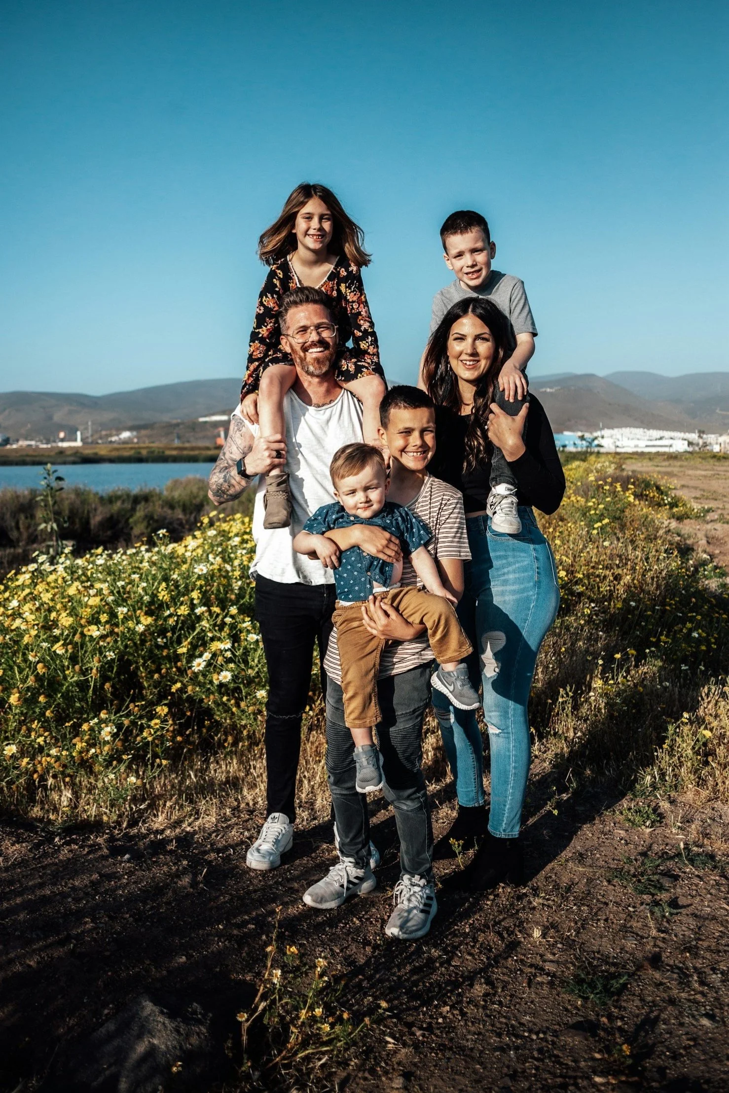 A multi-generational family of seven posing outdoors in a field of yellow flowers with mountains and a lake in the background on a sunny day.
