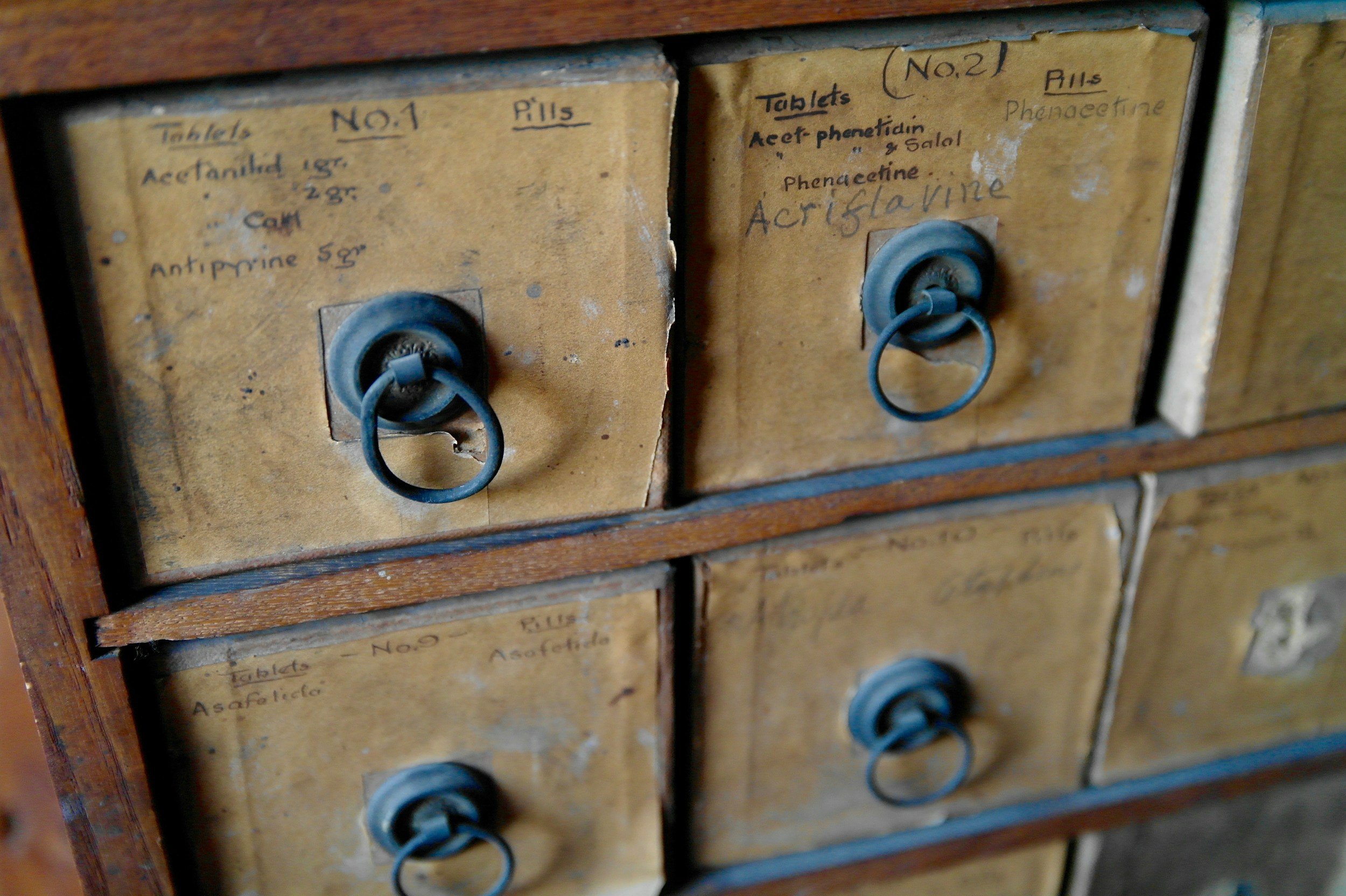 Antique wooden apothecary drawers with faded labels and round metal handles, showing names of old medications like Acetanilid and Phenacetin.