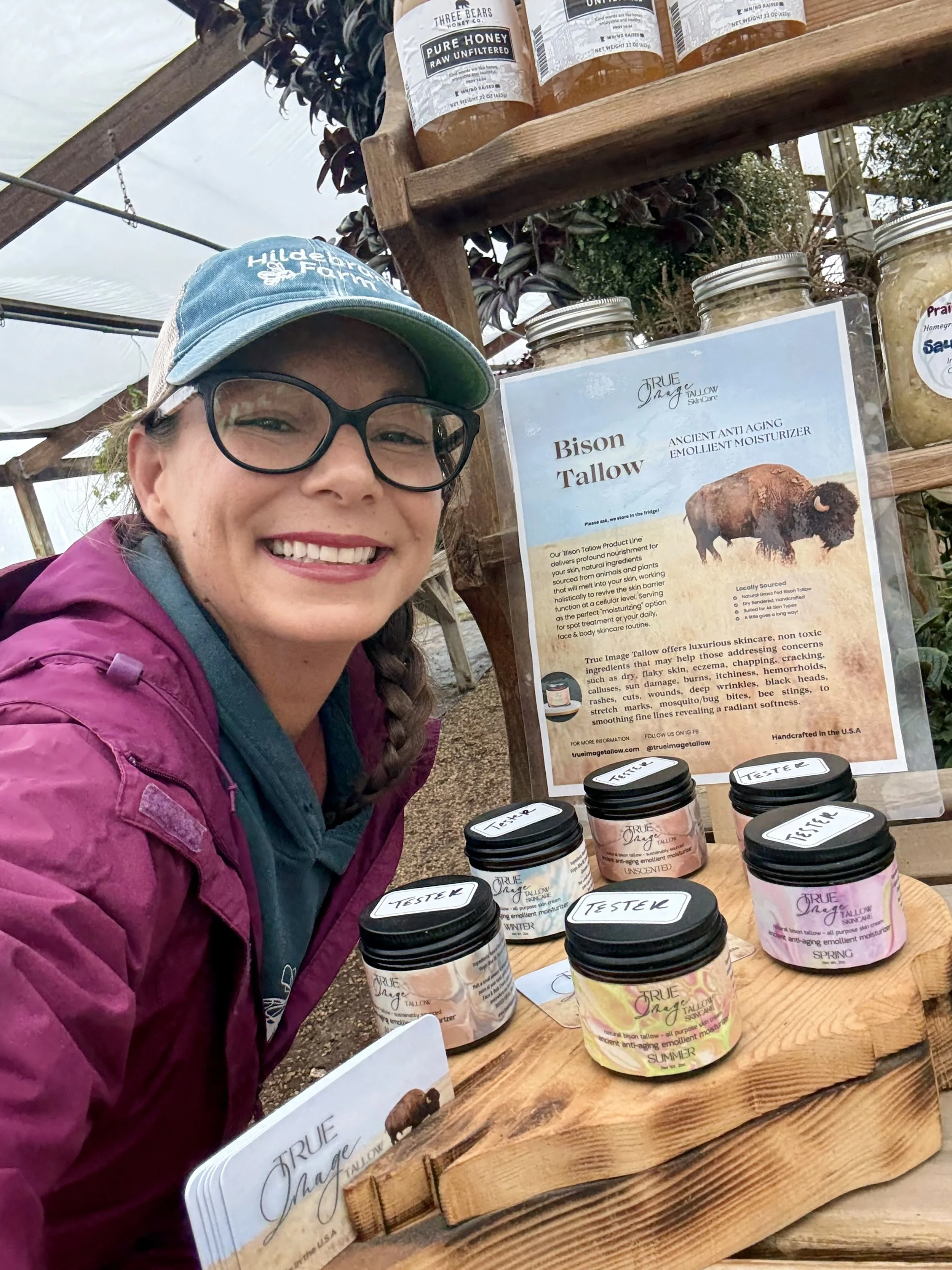 Woman smiling at a display of skincare products at an outdoor event. The display includes jars labeled 'Tester' containing various creams. A poster features an image of a bison and information about Tallow-based skincare.
