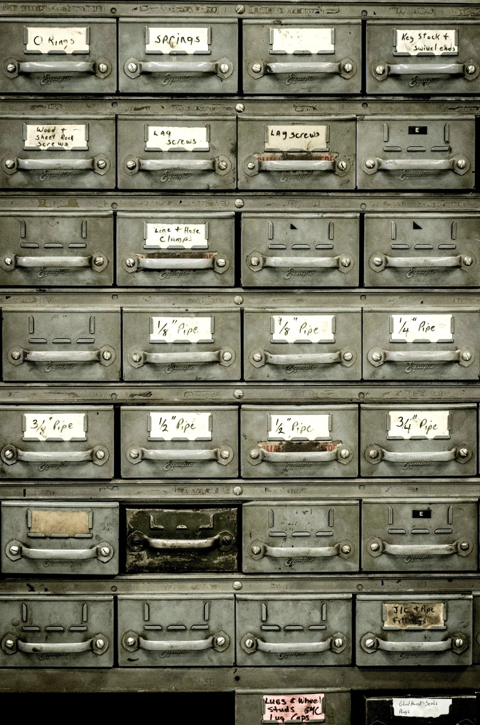 Vintage metal storage cabinet with labeled drawers for hardware items like springs, screws, and pipe fittings.