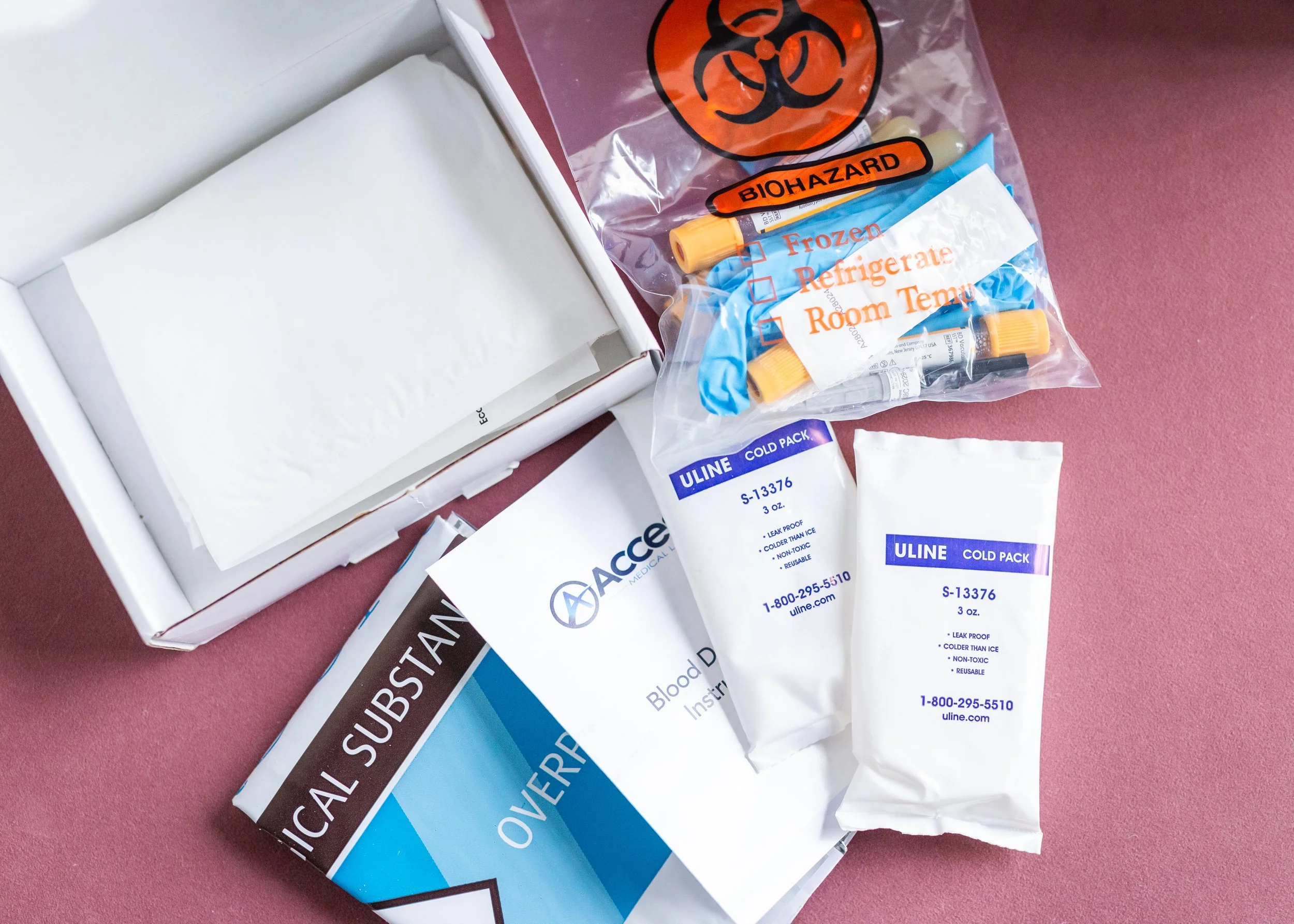Medical testing supplies including ice packs, a biohazard bag, blood collection instructions, and a box of sterile swabs on a pink surface.