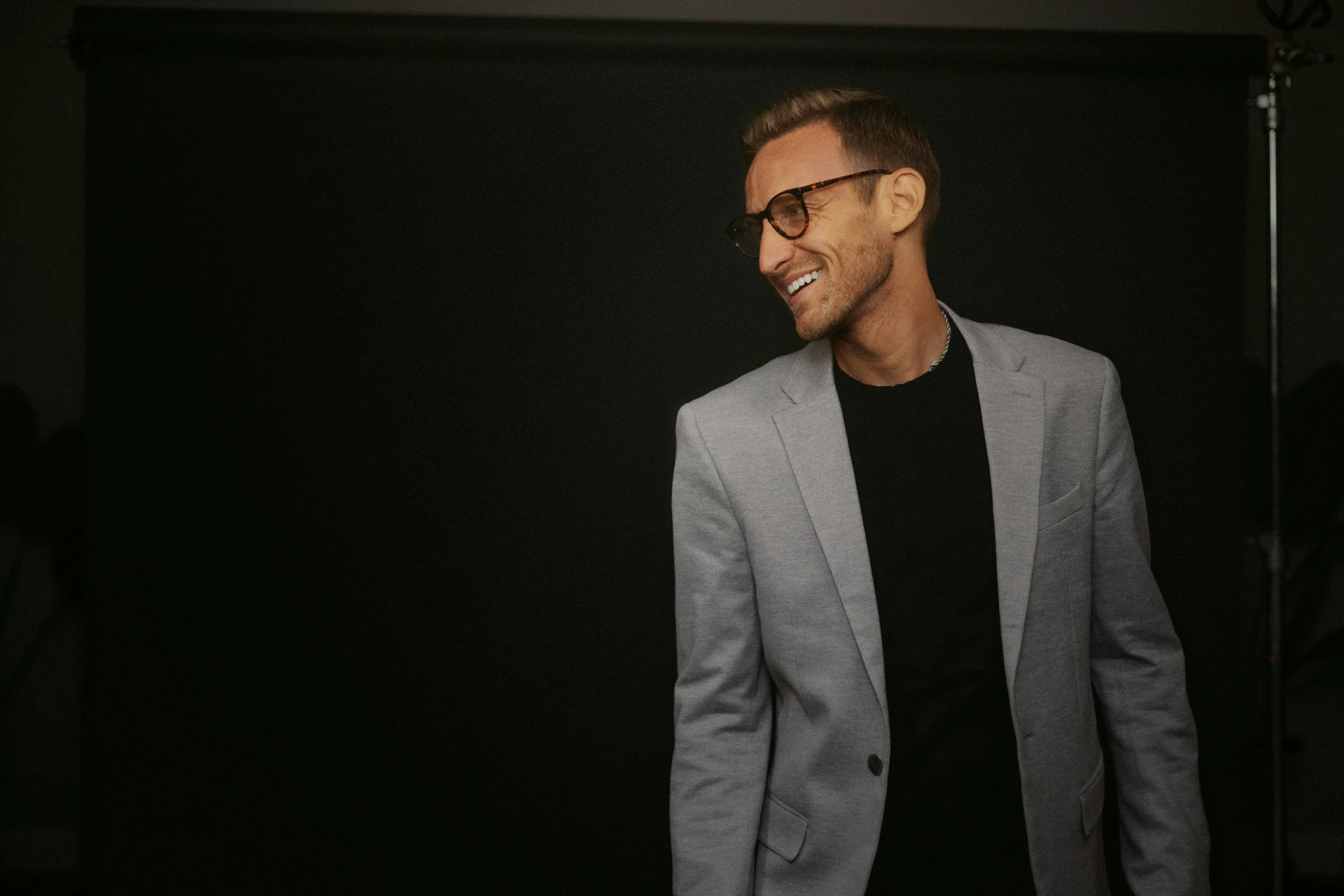 A man in glasses and a gray blazer smiling and looking to his left, standing against a dark background.