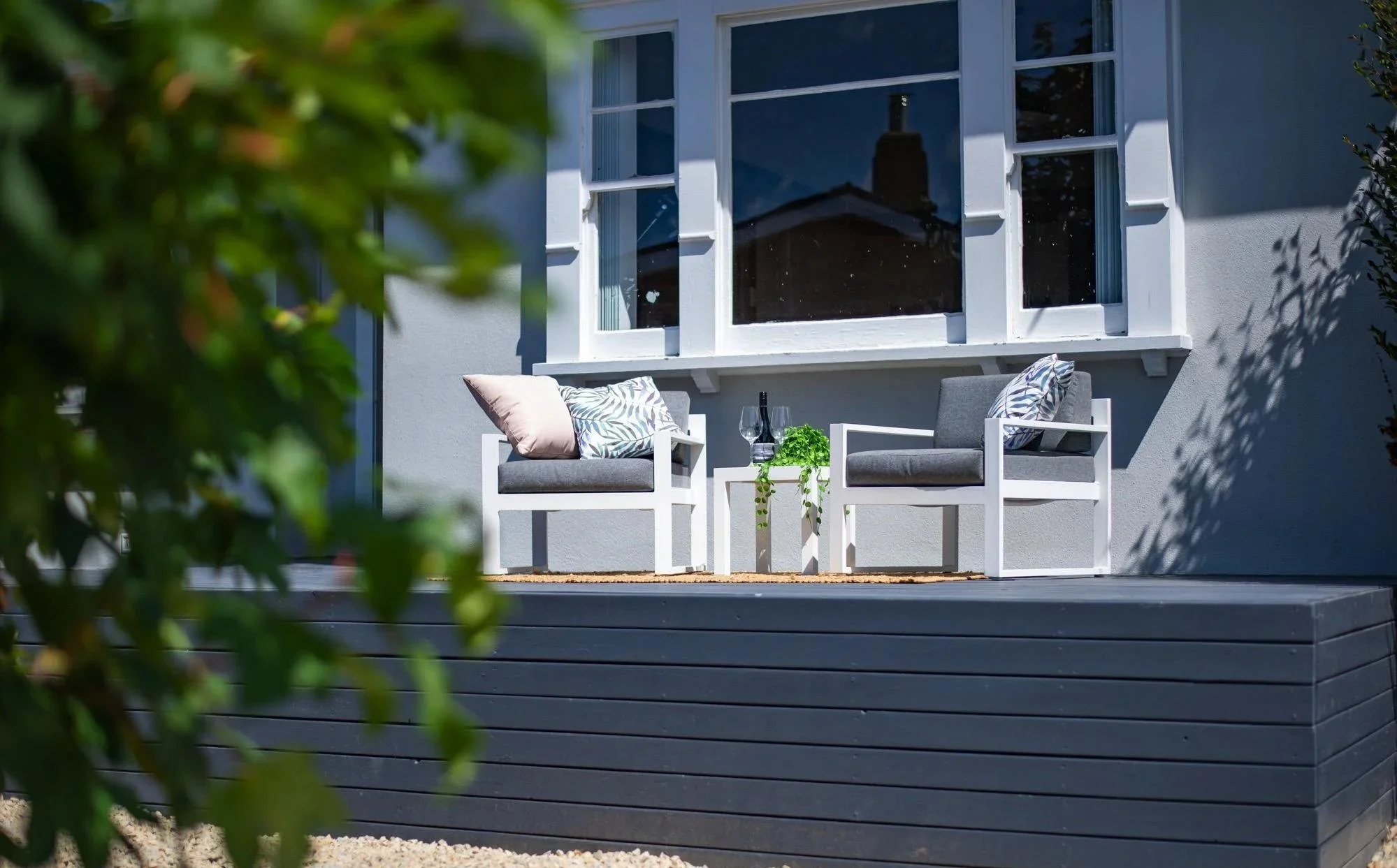 Outdoor patio with two gray armchairs, pillows, a small table with a plant, wine glasses, and a bottle, set against a house wall with a large window.