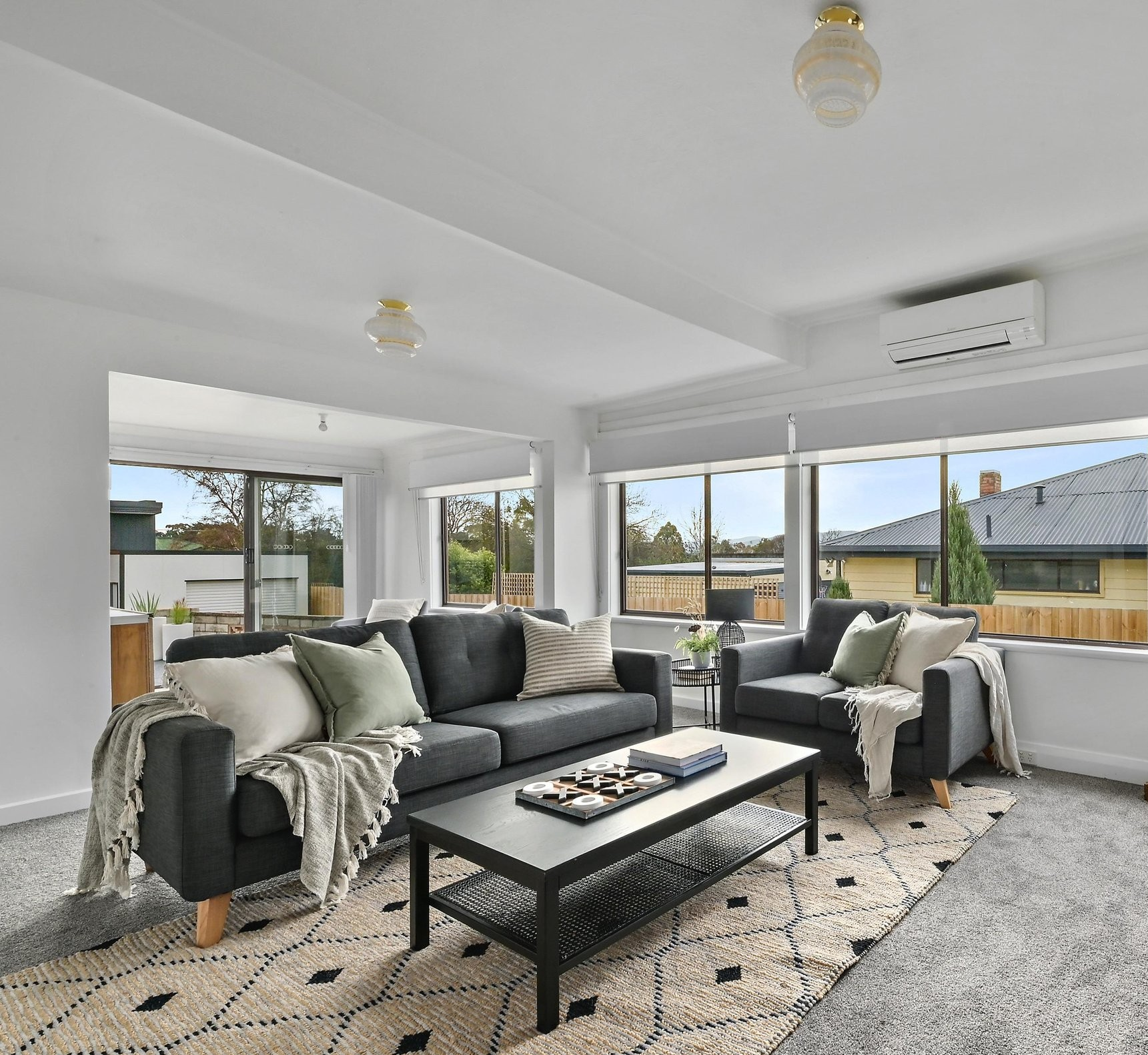 Living room with two dark gray sofas, light-colored throw blankets and pillows, a black coffee table with books and decorative items, large windows with blinds showing an outdoor view, and a patterned rug on gray carpet flooring.