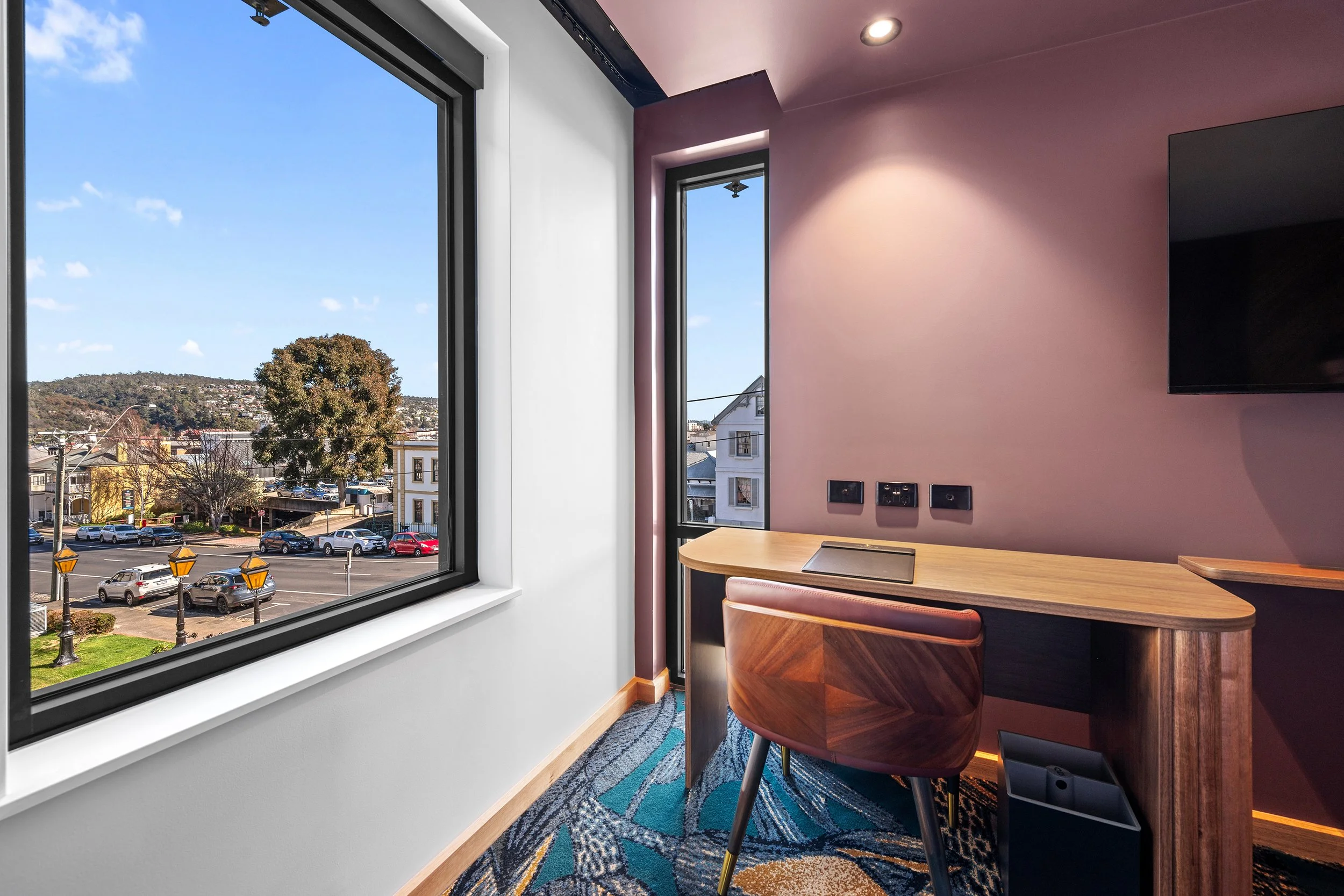 View from a small office with pink walls, a wooden desk, a leather chair, and a flat-screen TV on the wall. Large windows show a parking lot and trees outside.