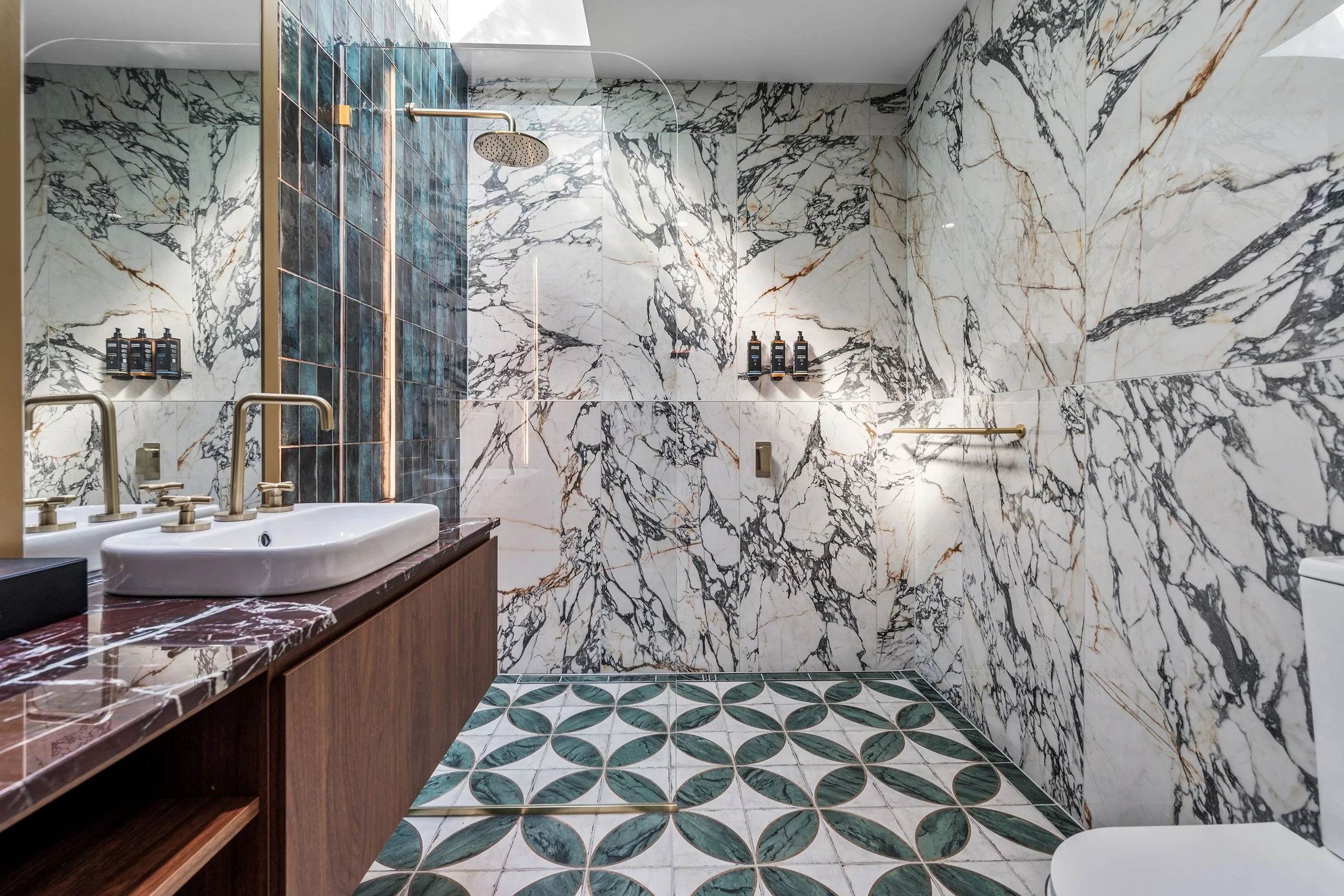 A luxurious bathroom with marble walls, a wooden vanity with a white sink, a large mirror, a shower area with a rain shower head, and decorative tiled flooring with circular green and white patterns.