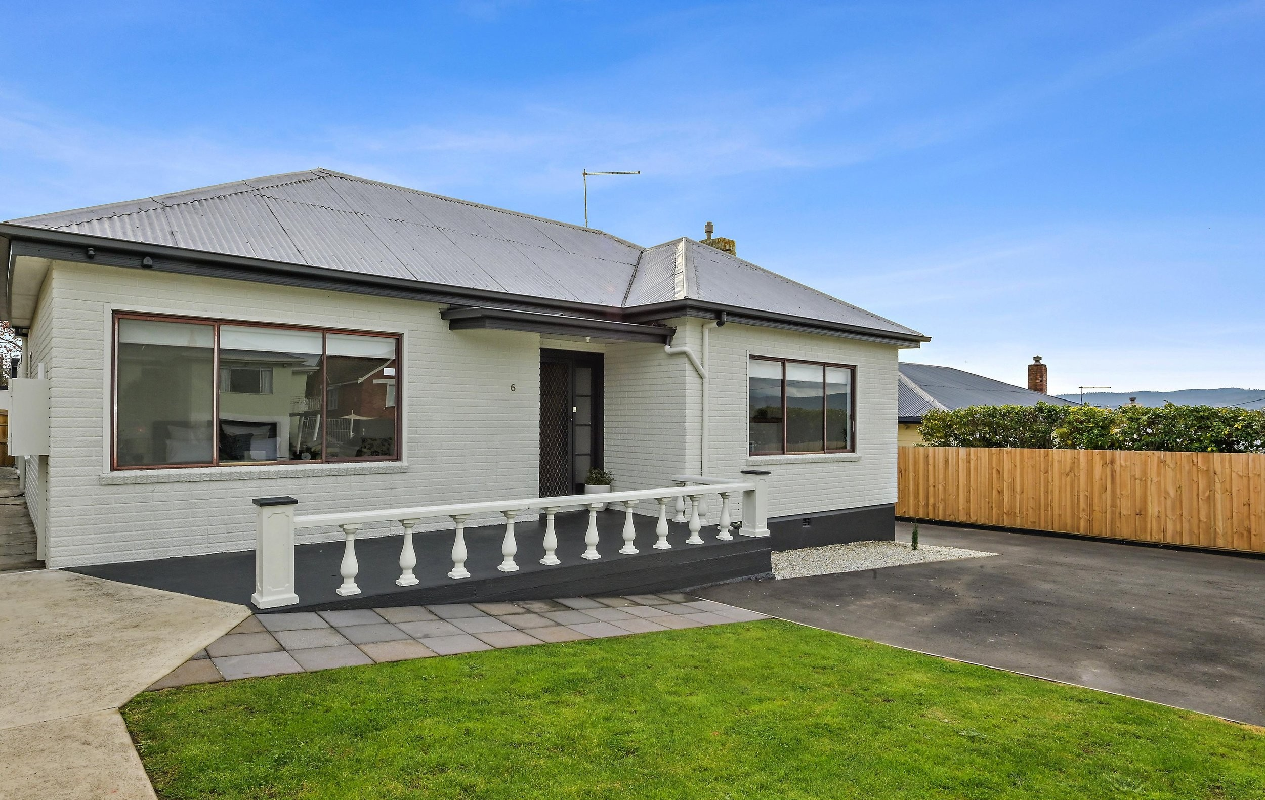 White house with a black door, large front windows, a small porch with white railing, a driveway, a grassy yard, and a wooden fence in the background under a blue sky.