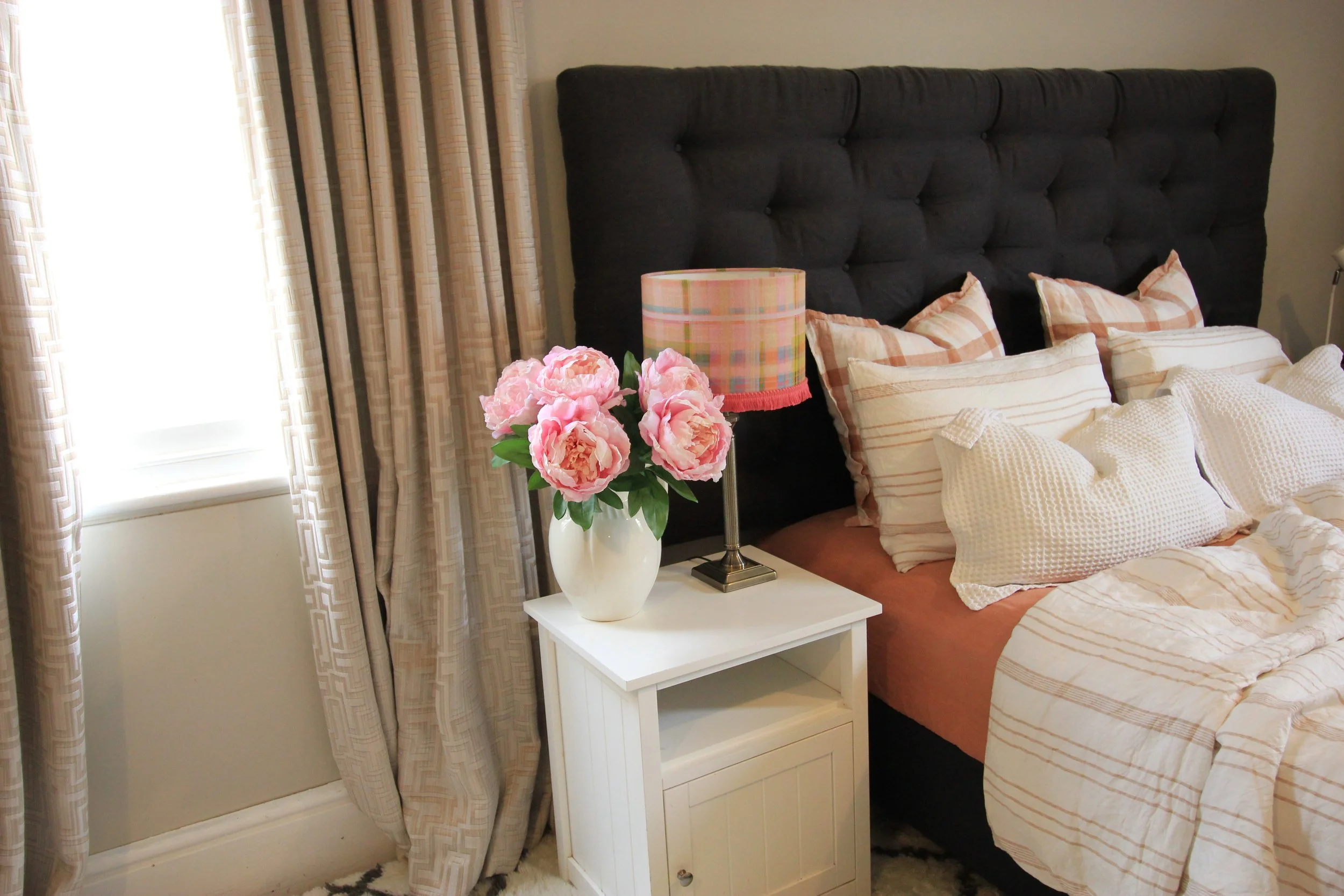 A cozy bedroom with a white nightstand holding a vase of pink peonies and a pink plaid lampshade. The bed has a dark tufted headboard and striped pink and white bedding, with multiple pillows. Curtains frame the window on the left.
