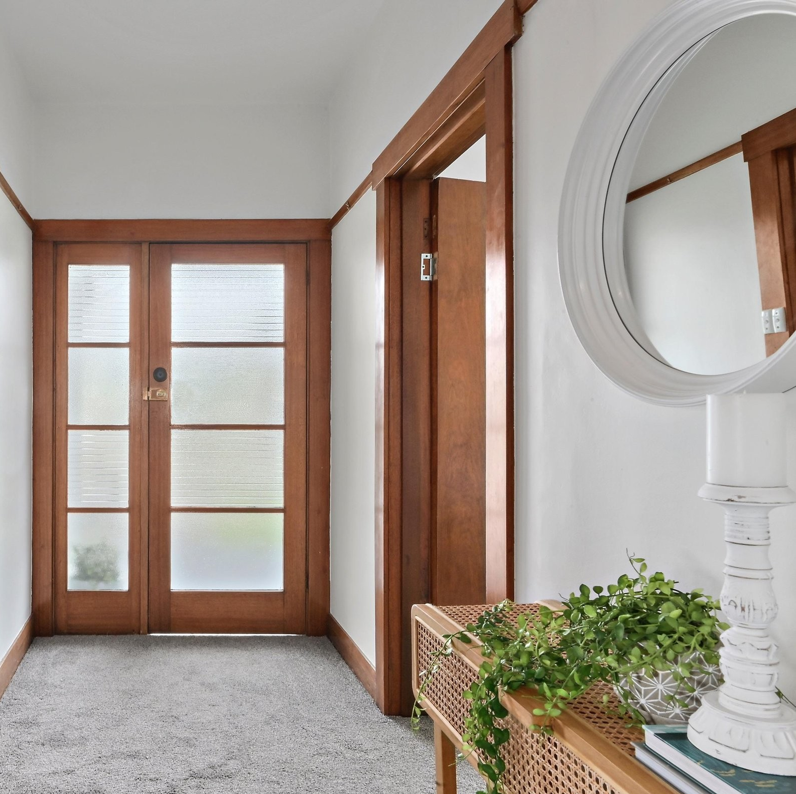 Entryway with wooden-framed glass double doors, a white wall, a round mirror, a wooden console table with potted plant, candlestick, and books.