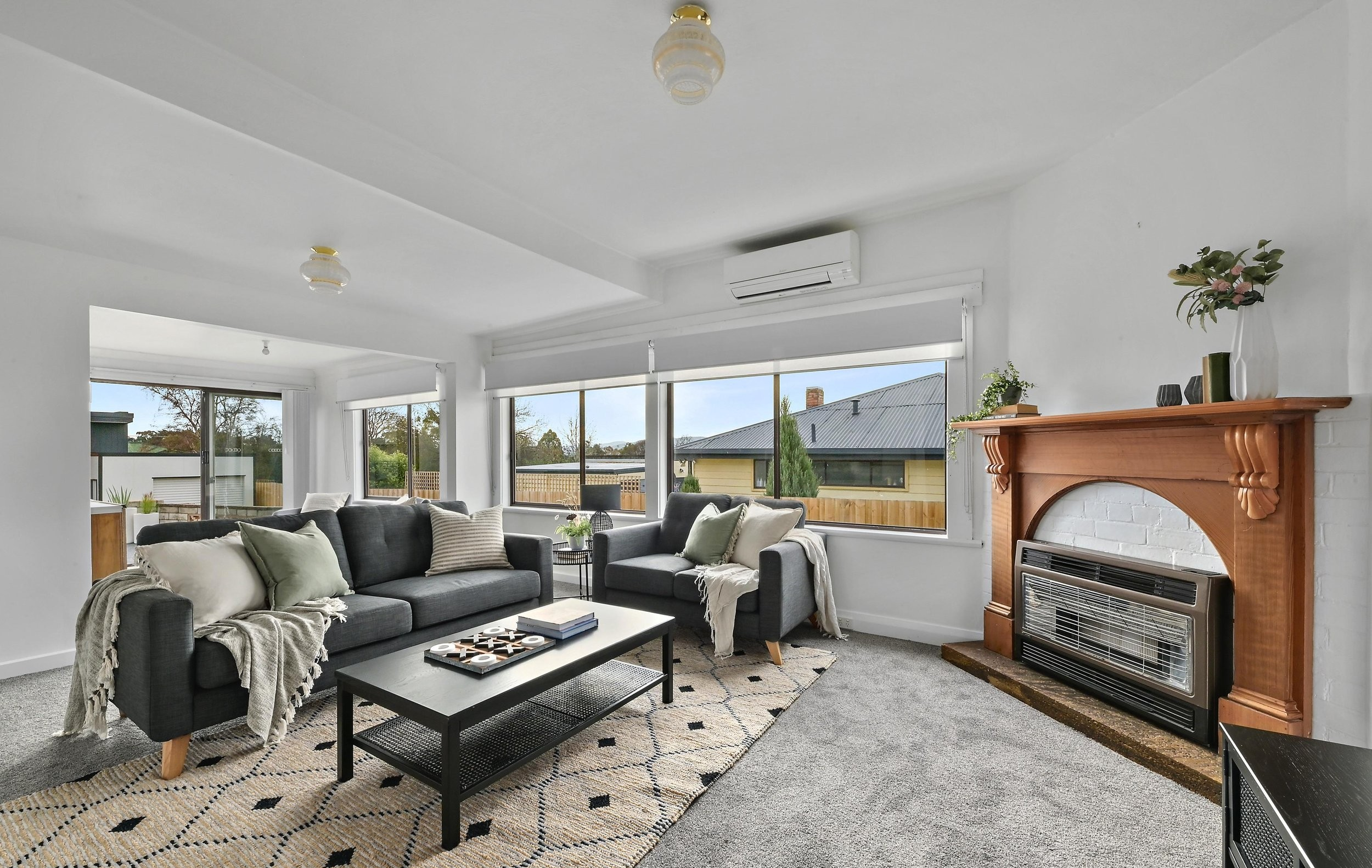 Bright living room with gray sofas, throw pillows, and a patterned rug. Large windows with view of neighboring houses. Fireplace with wooden mantel and decorative items, wall-mounted air conditioning unit.