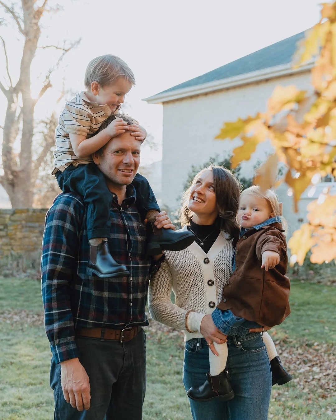 A family of four outdoors in autumn, with kids sitting on parents' shoulders, smiling and enjoying the moment.