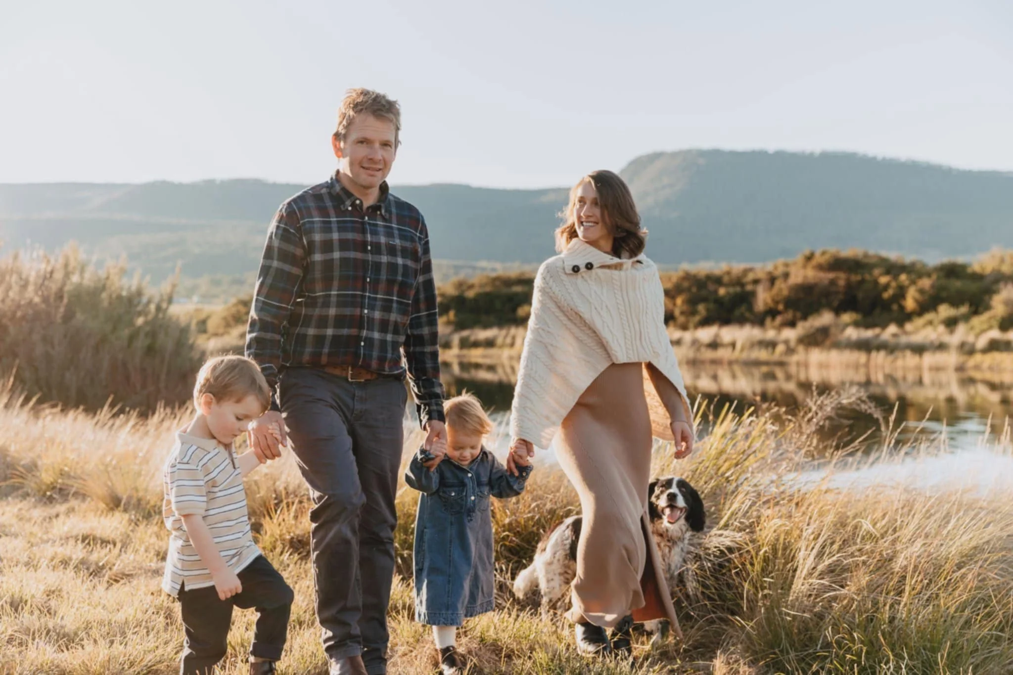 A family of four walking outdoors near a lake during sunset, with a black and white dog, surrounded by tall grass and mountains in the background.
