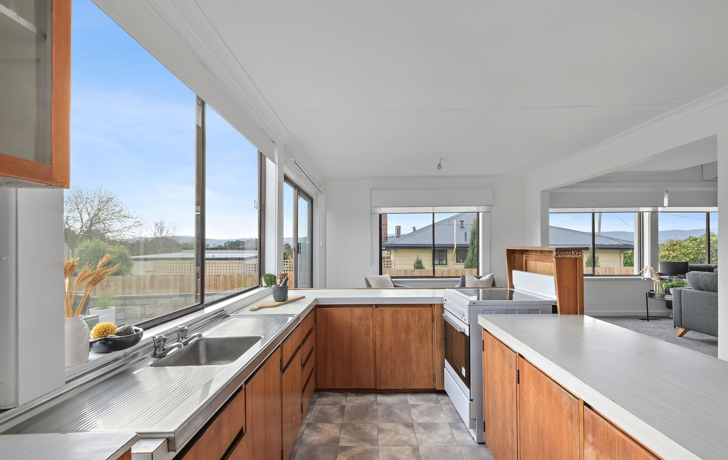 Kitchen with large windows, wooden cabinets, stainless steel sink, stove, and a view of a backyard with neighboring houses and trees.