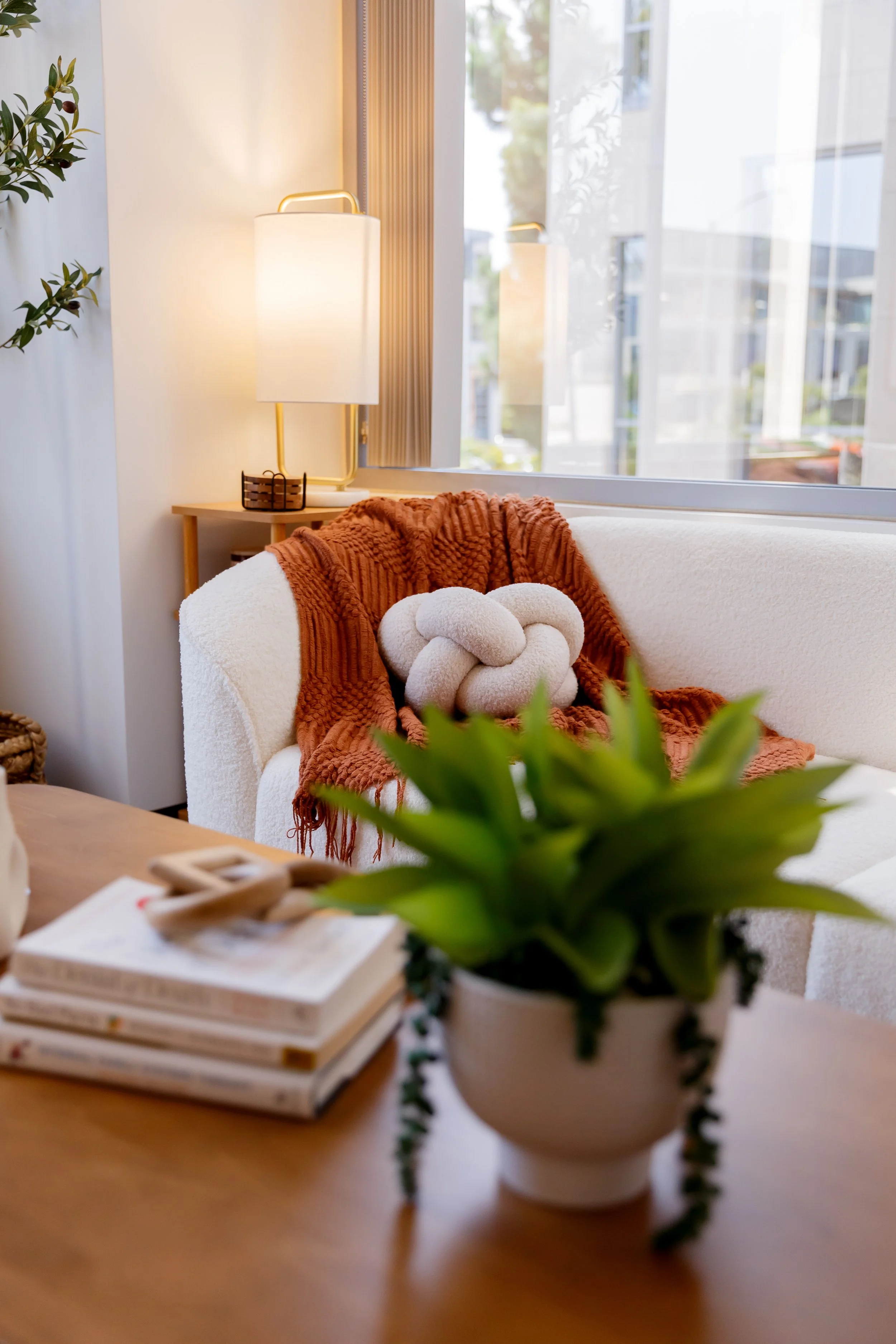 A cozy living room corner with a white sofa, a rust-colored blanket, a knot-shaped cushion, a wooden side table with a lamp, a large window with sheer curtains, a potted plant on a wooden table, and a stack of magazines on the table.