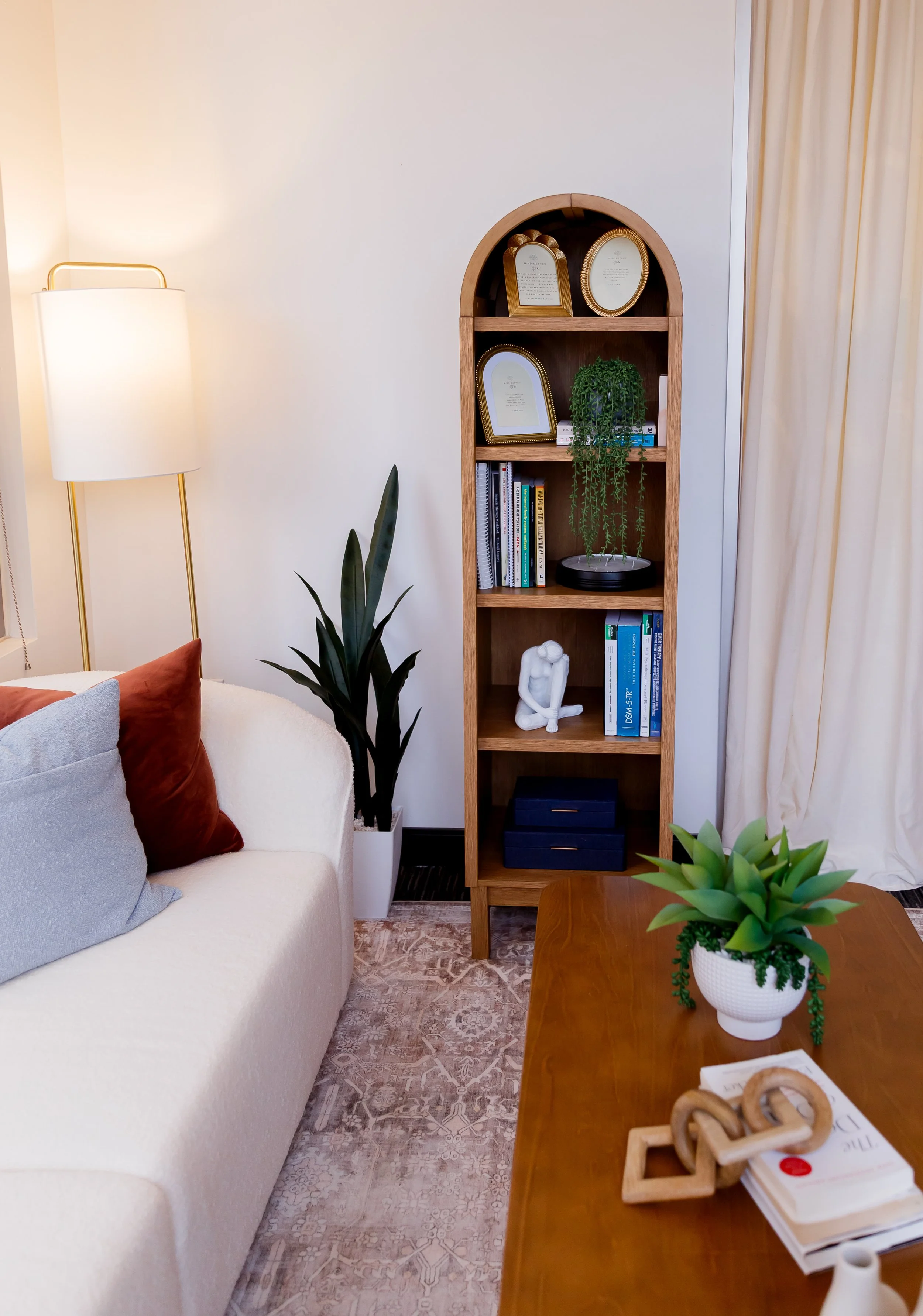 A cozy living room with a white sofa featuring gray and red pillows, a wooden bookshelf with plants, books, and decorative items, a potted plant on a wooden table, and a tall floor lamp beside the sofa. The room has light-colored walls and curtains, with patterned carpeting.