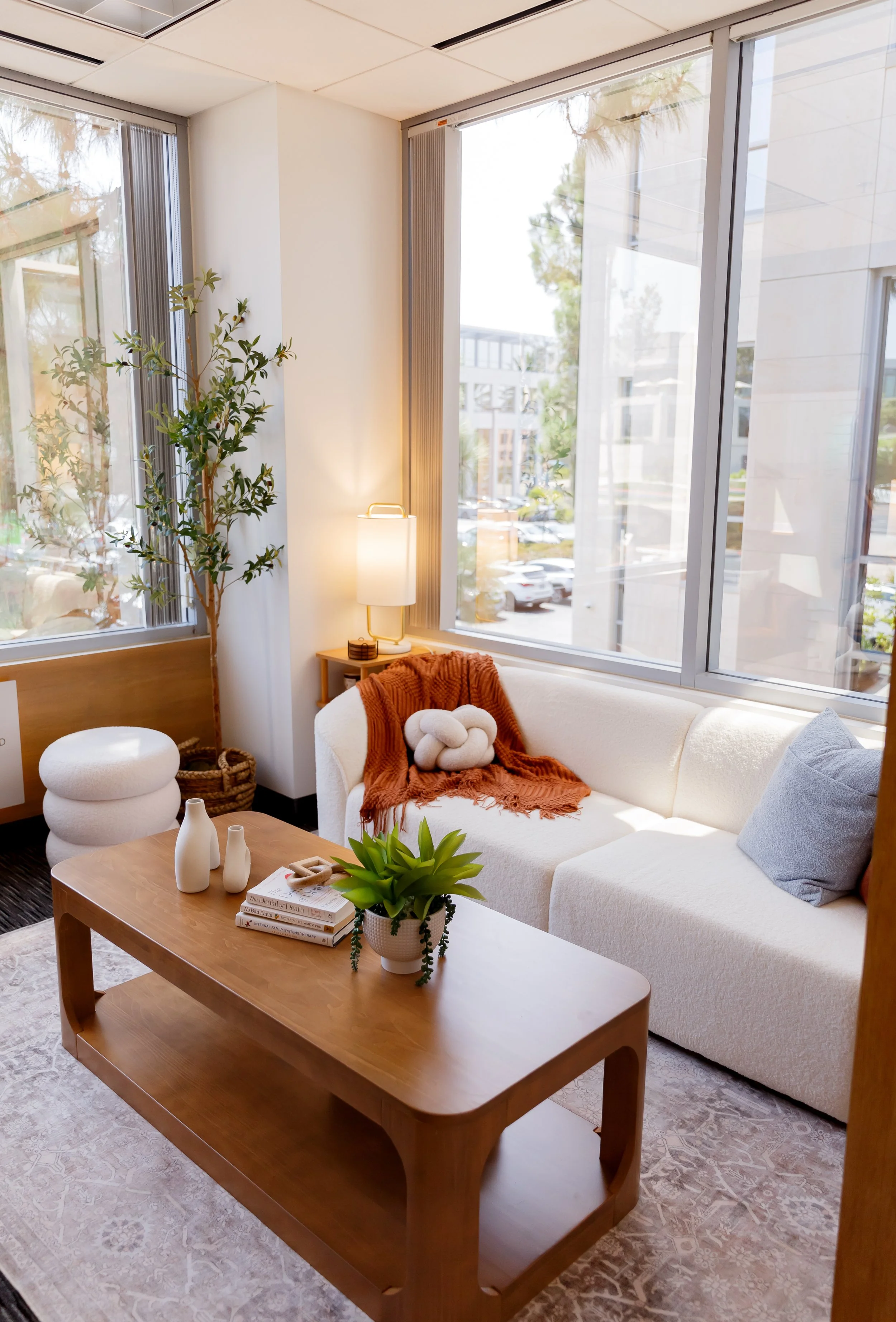 A cozy living room with a large white sectional sofa near floor-to-ceiling windows. The sofa has two pillows, one gray and one blue, and a rust-colored throw blanket with knot details. A wooden coffee table holds a potted green plant, two white vases, a book, and a small sculptural object. A tall lamp with a white shade is next to a side table. A potted plant and woven basket are in the corner. Outside, a parking lot and modern buildings are visible through the windows.