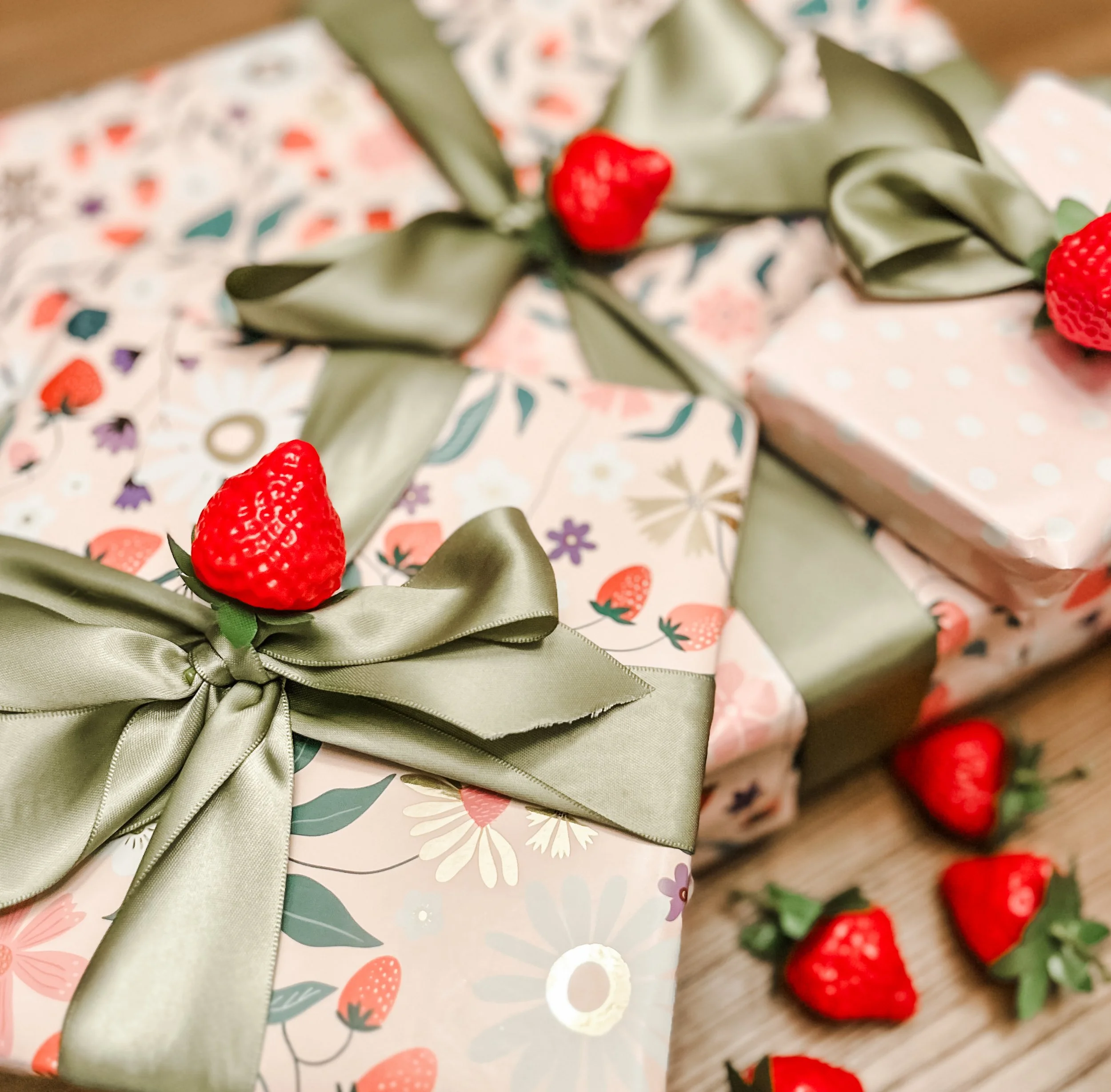 Three wrapped gifts in pink paper that has florals and strawberries on it with sage green satin ribbons and strawberry decor attached