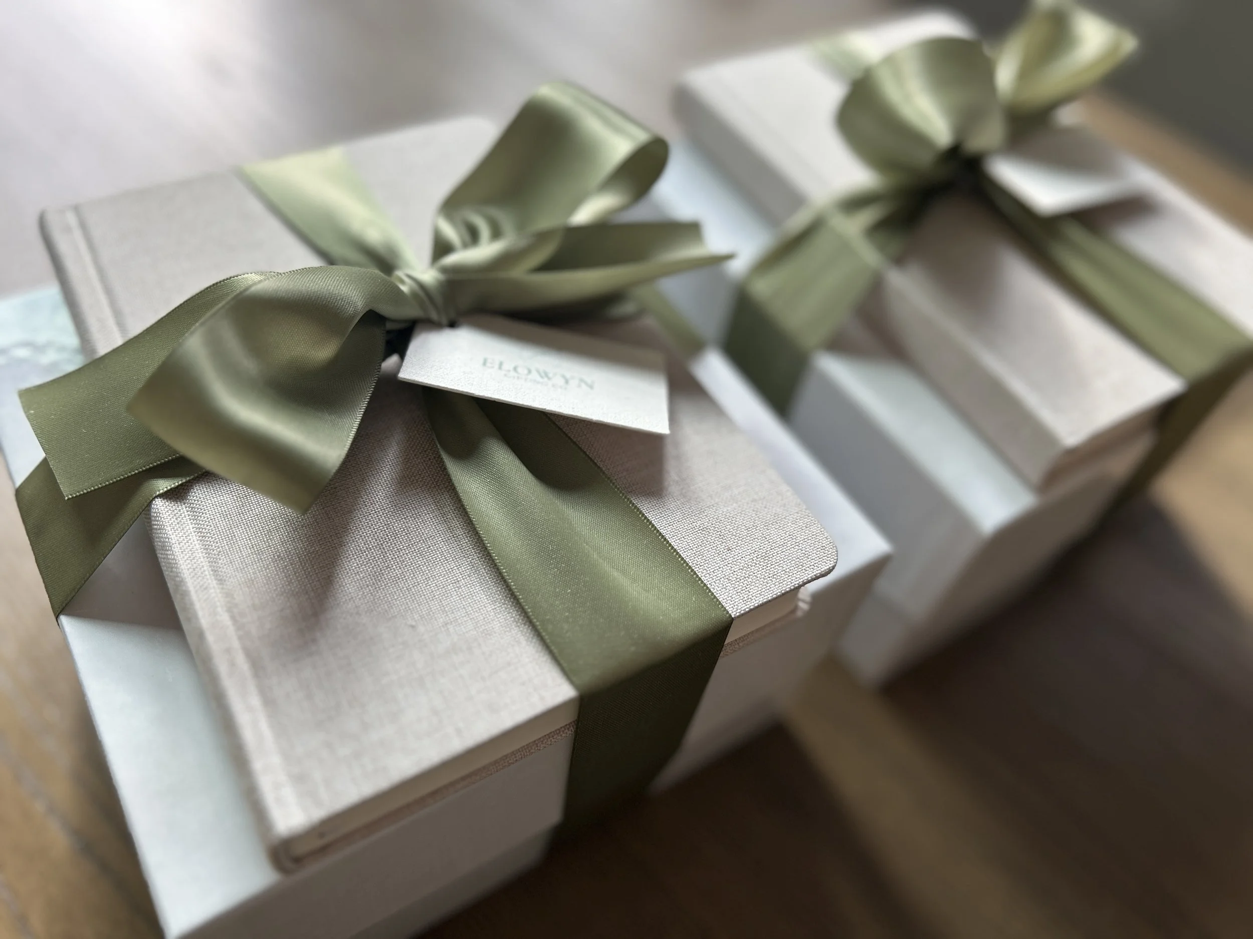 Two white gift boxes with a journal tied to the top with a gift tag and sage green satin ribbons