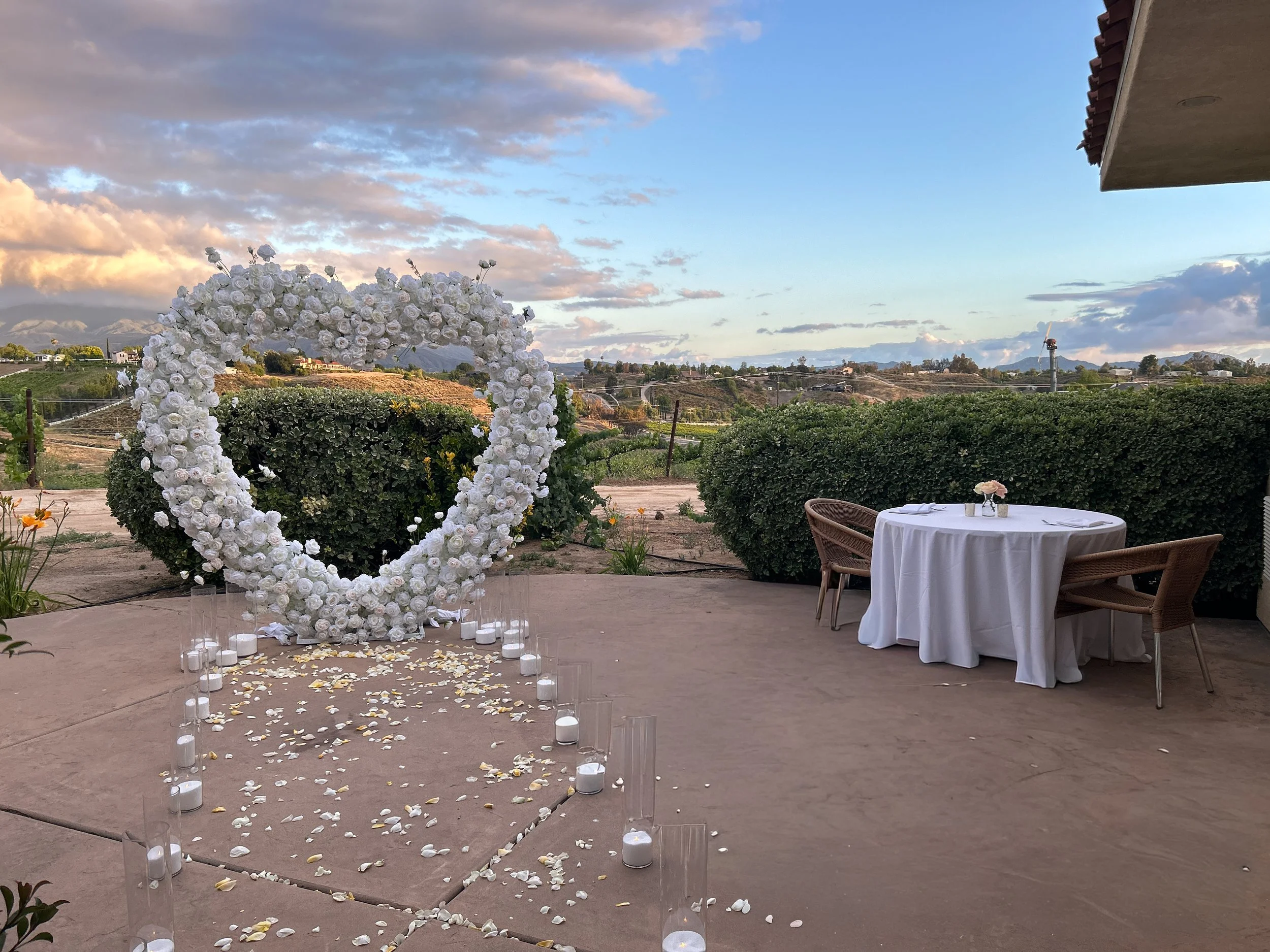 heart arch proposal with a neon will you marry me sign set up with canldes and rose petals next to a private dining table on a personal villa patio
