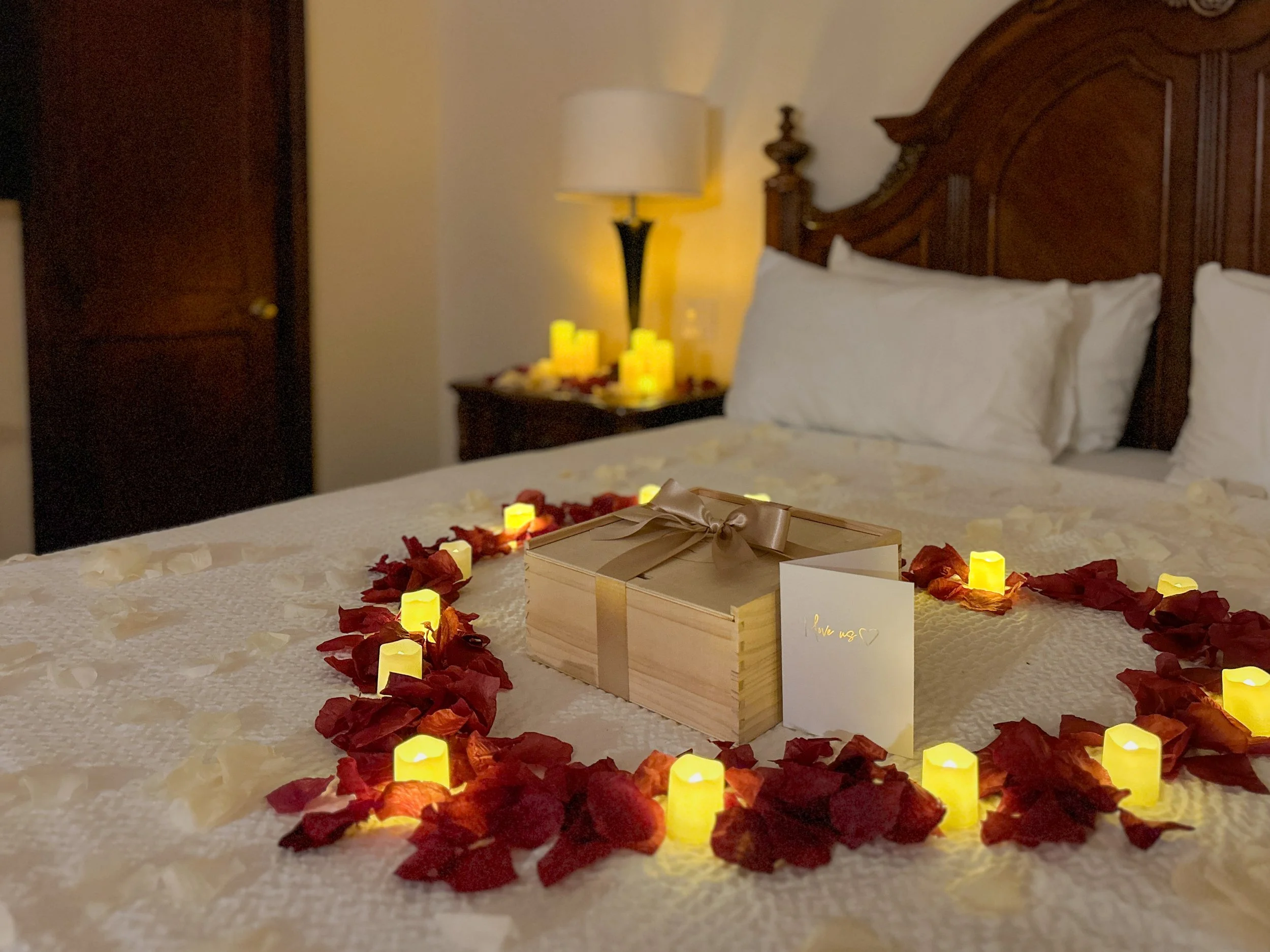 a hotel bed covered with a rose petal heart and candles with an engraved wooden keepsake box and a card in the middle