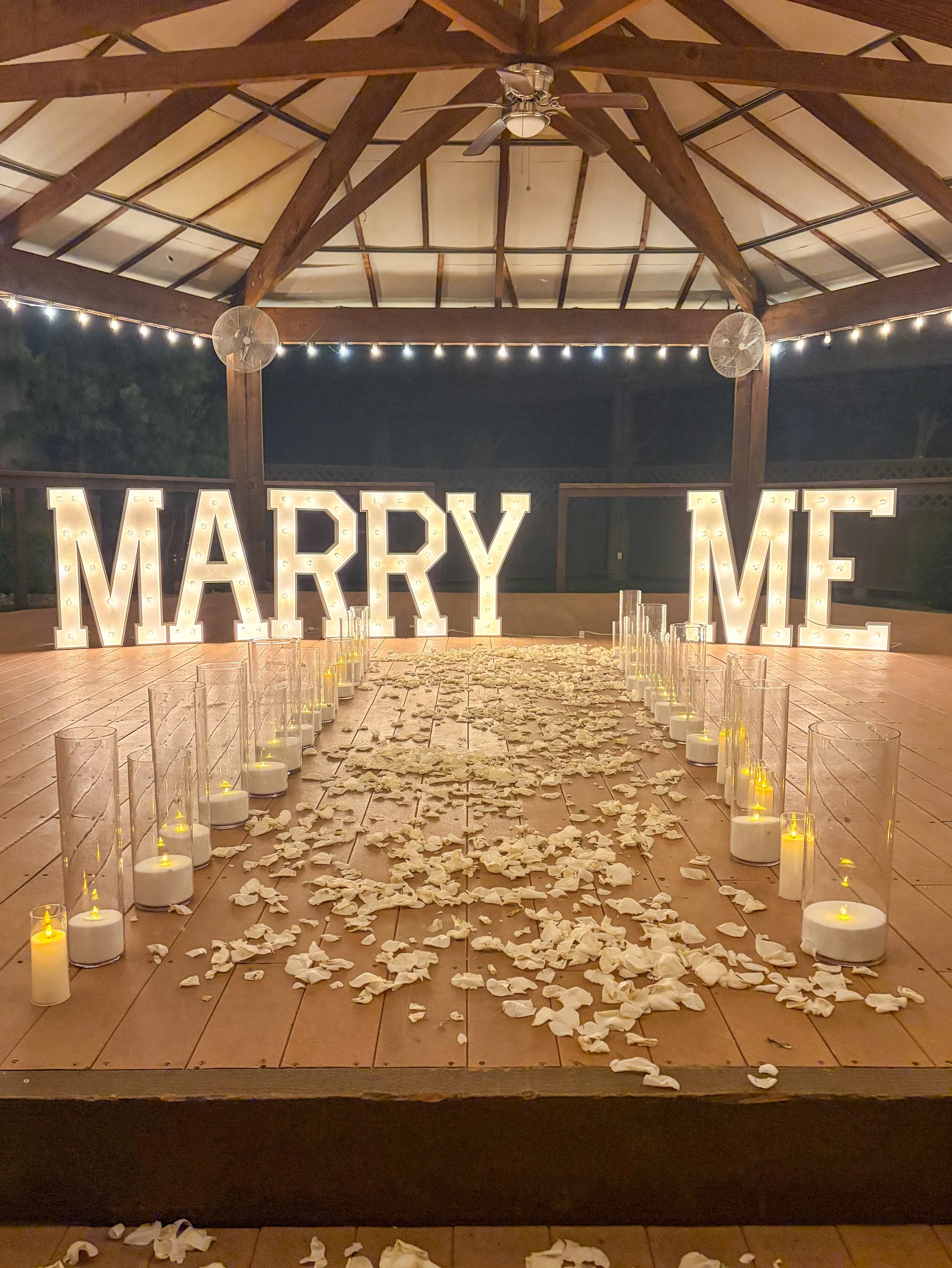 Large lit up marquee letters that say Marry Me with candles and rose petals