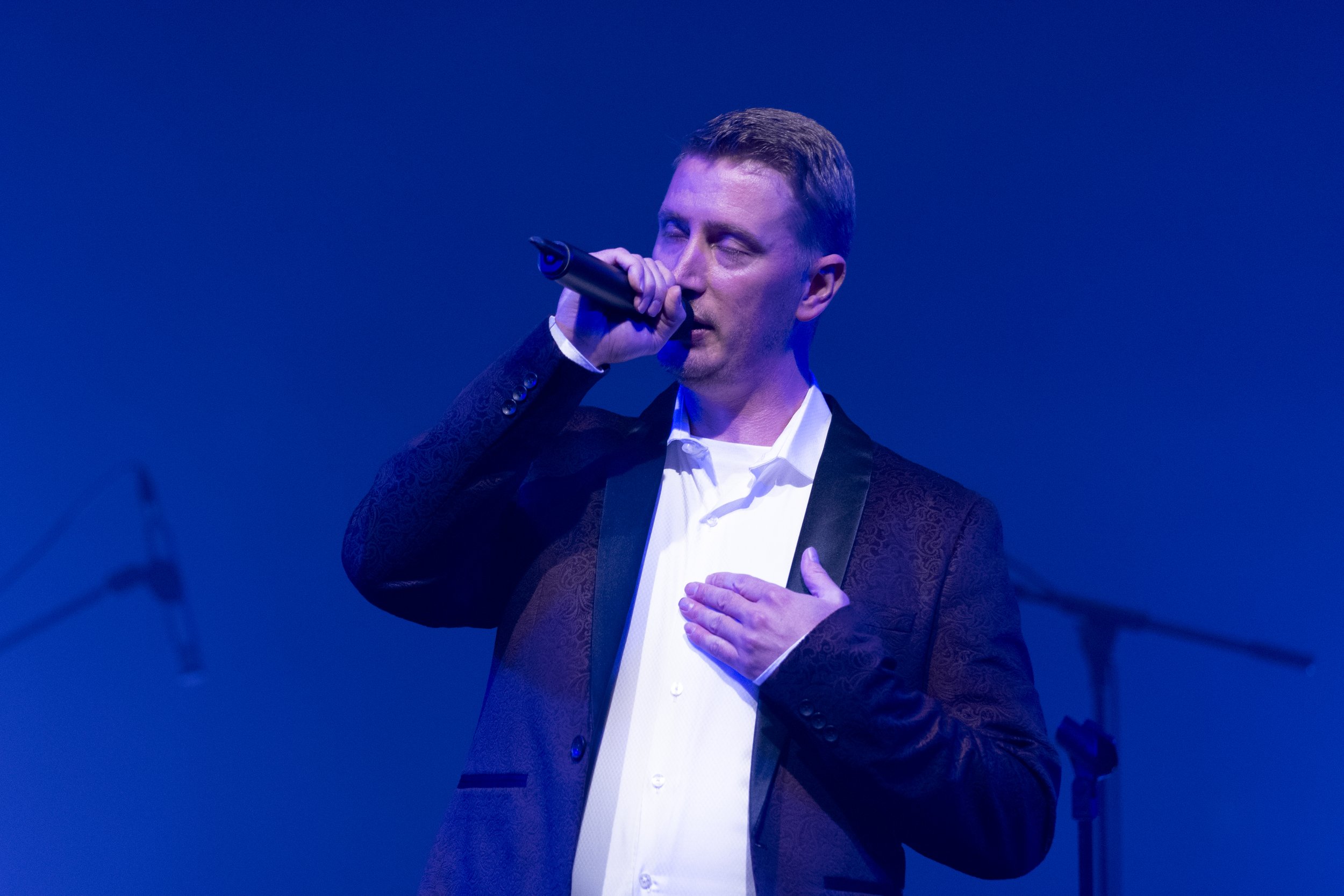 Man in suit singing into a microphone on stage with blue lighting.