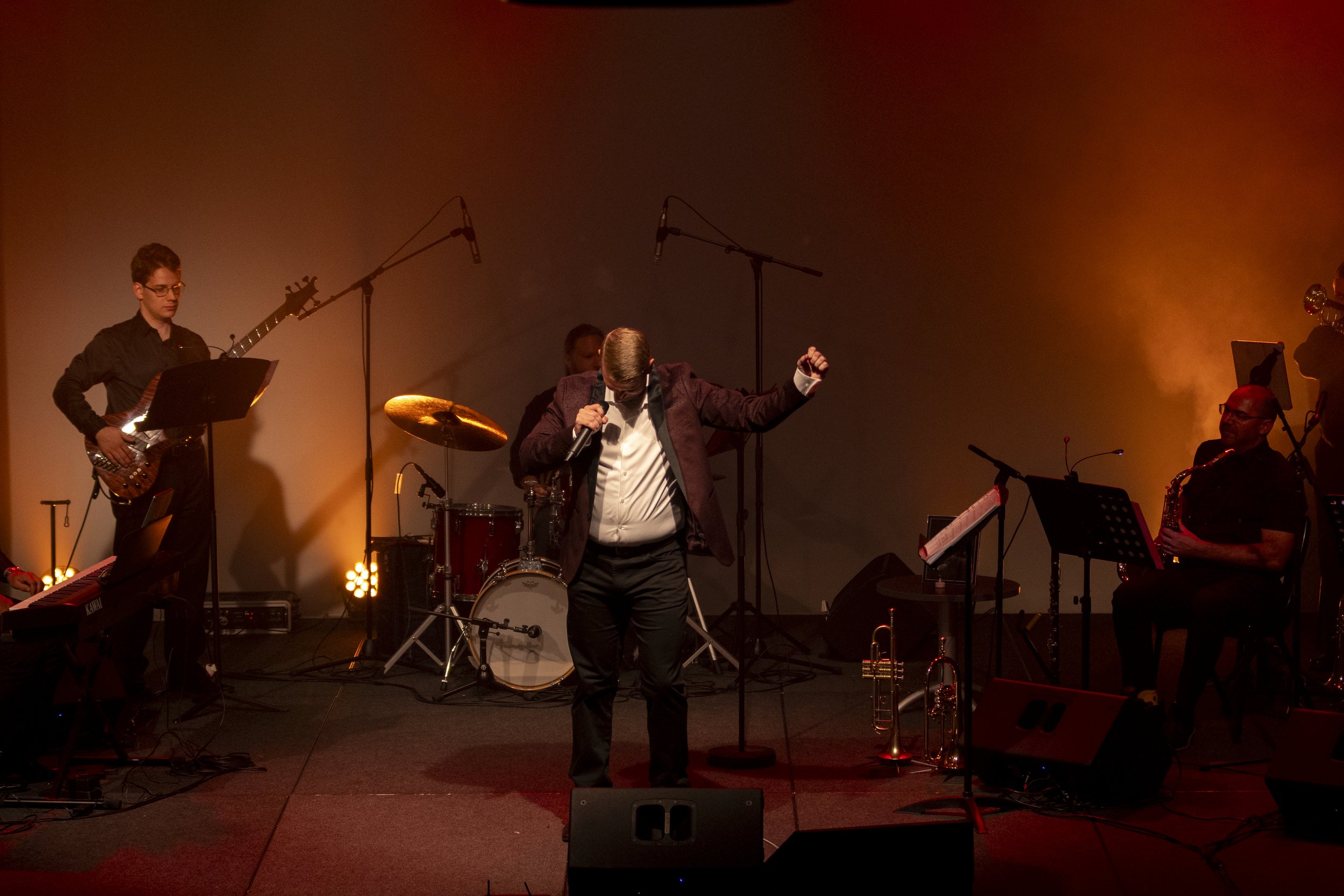 A live band performing on stage with a lead singer holding a microphone. Musicians play instruments including guitar, drums, and saxophone, under dramatic stage lighting.