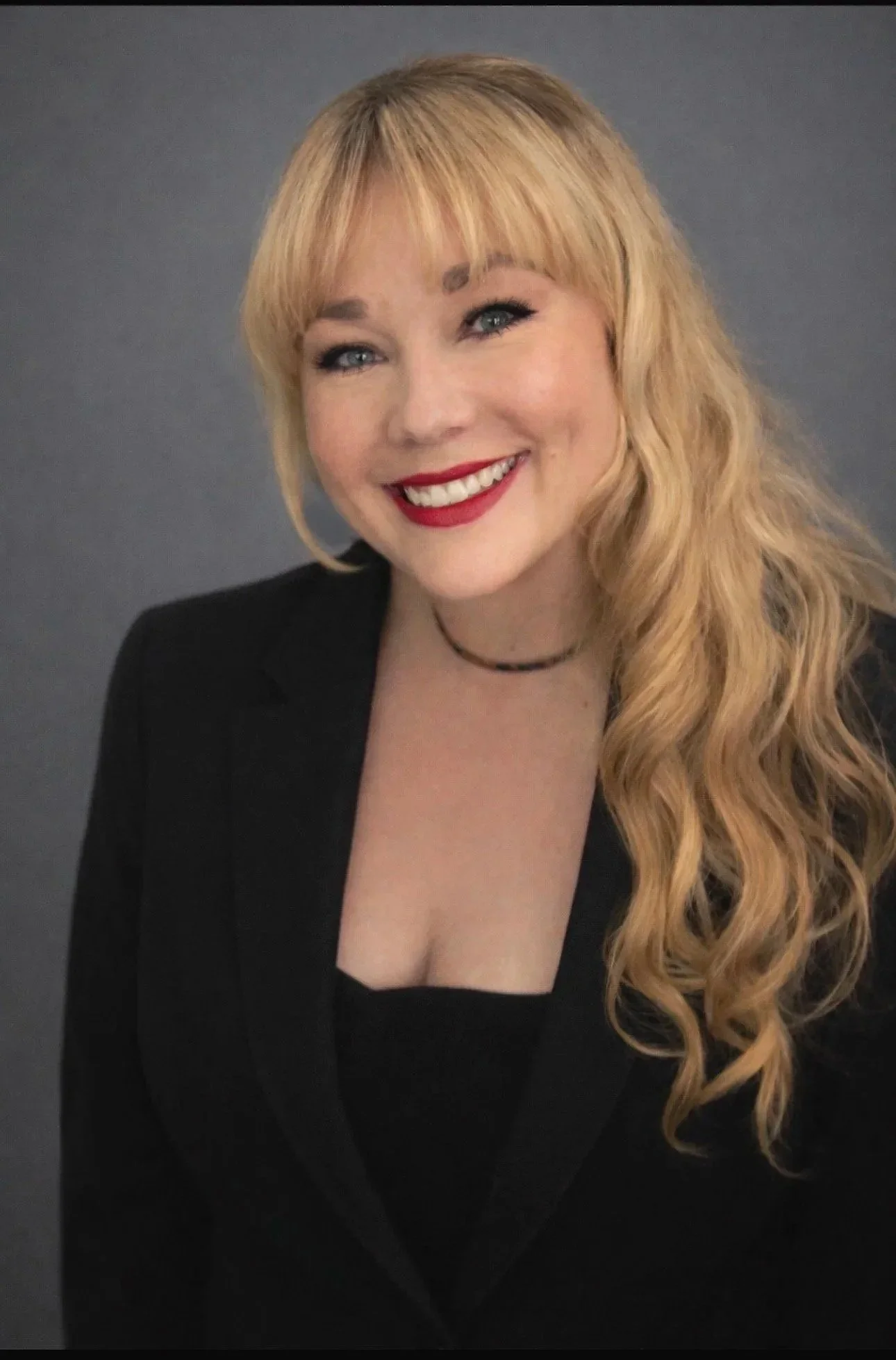 A woman with long wavy blonde hair wearing a black blazer and a choker necklace, smiling with red lipstick against a grey background.