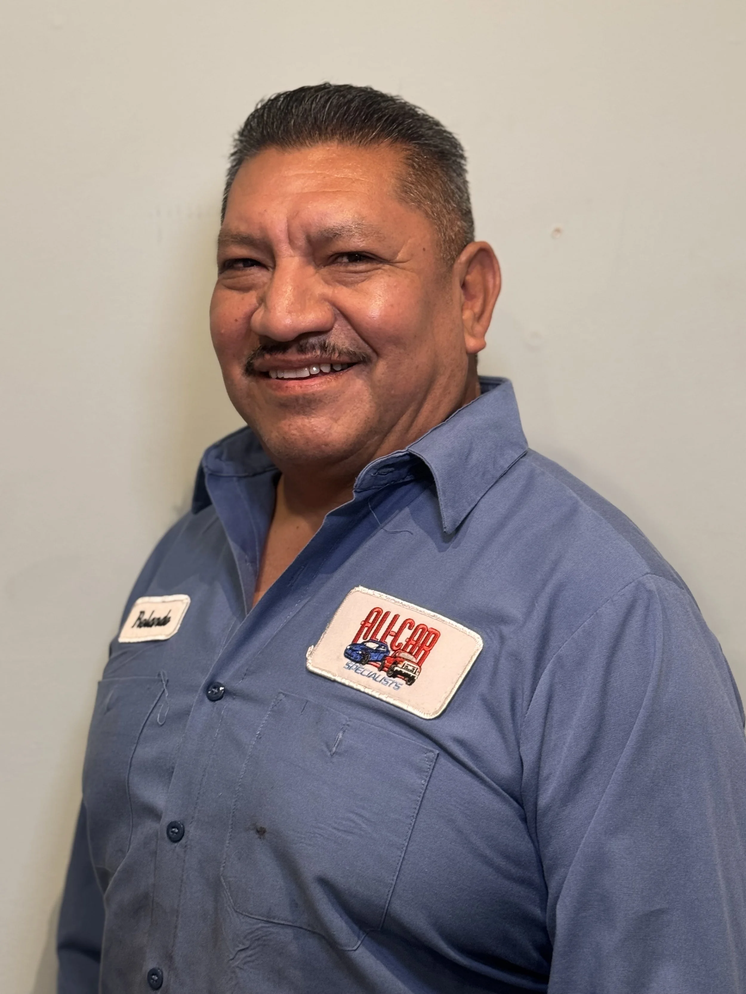 A man with a mustache and a blue work shirt. The Patch reads 'ALLCAR'. Standing in front of a plain wall.