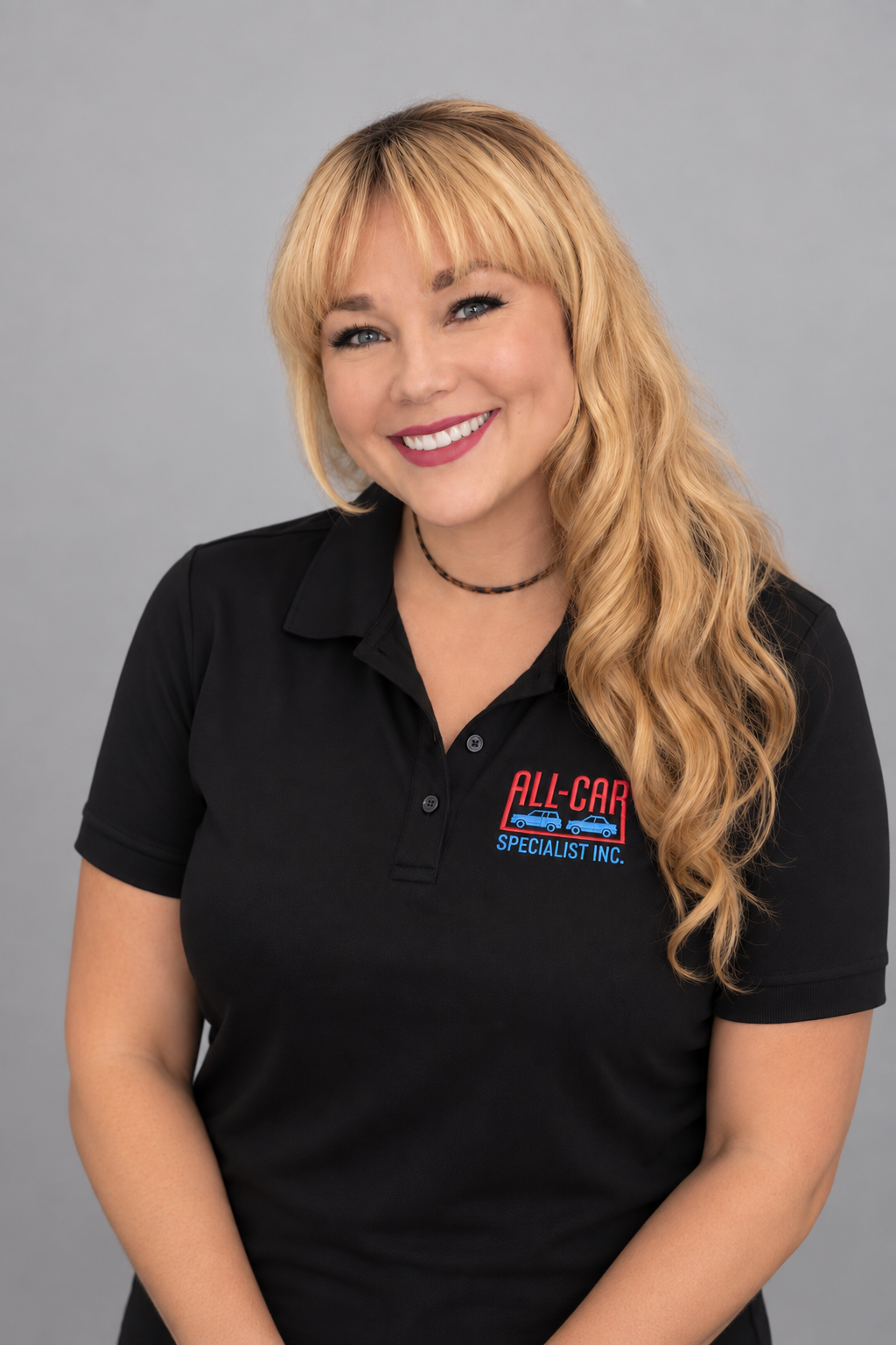 A woman with long wavy blonde hair wearing a Black polo with All- Car Specialist Embordered and a choker necklace, smiling with red lipstick against a grey background.