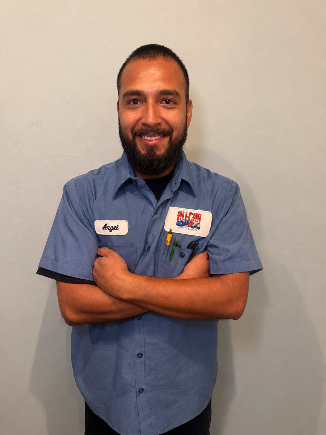 A man smiling, in a blue work shirt with a name tag stands in front of a plain light-colored wall.