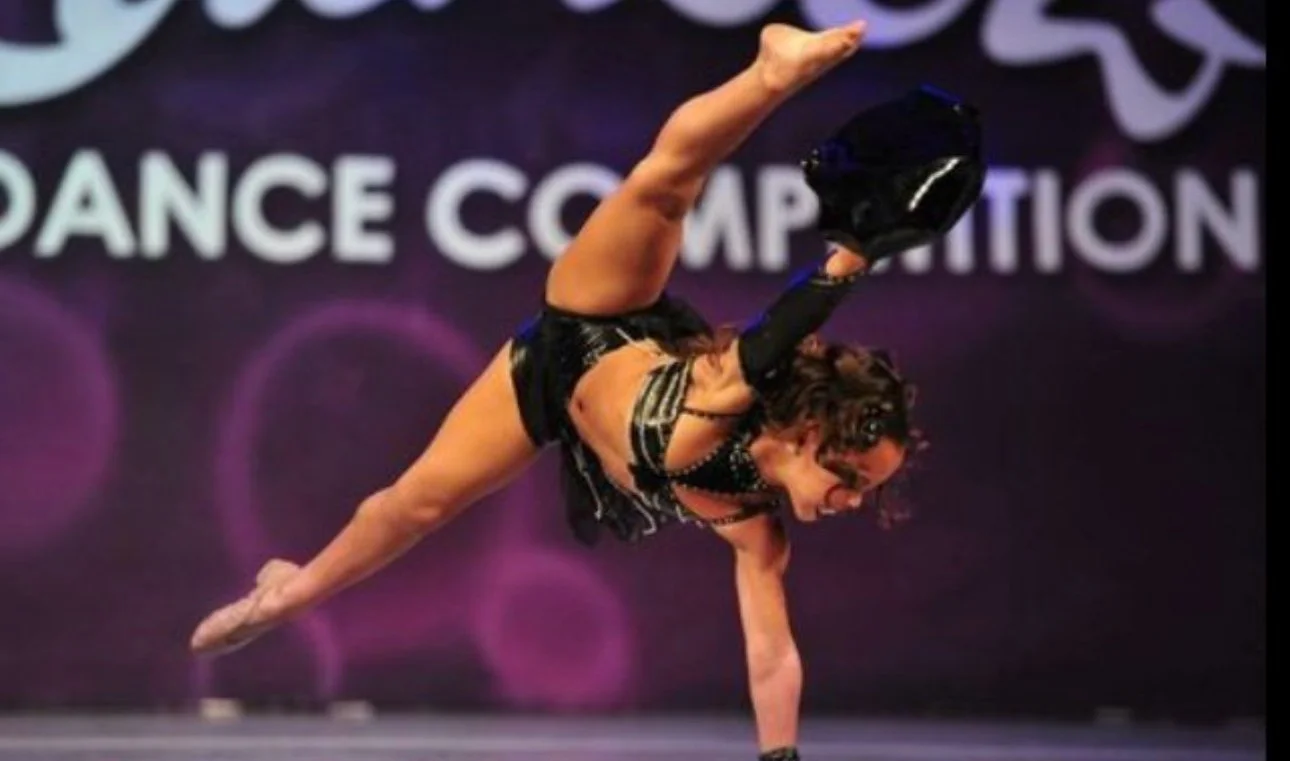 A female dancer in a black costume performing a handstand on stage with a backdrop that has the words 'Dance Competition'.