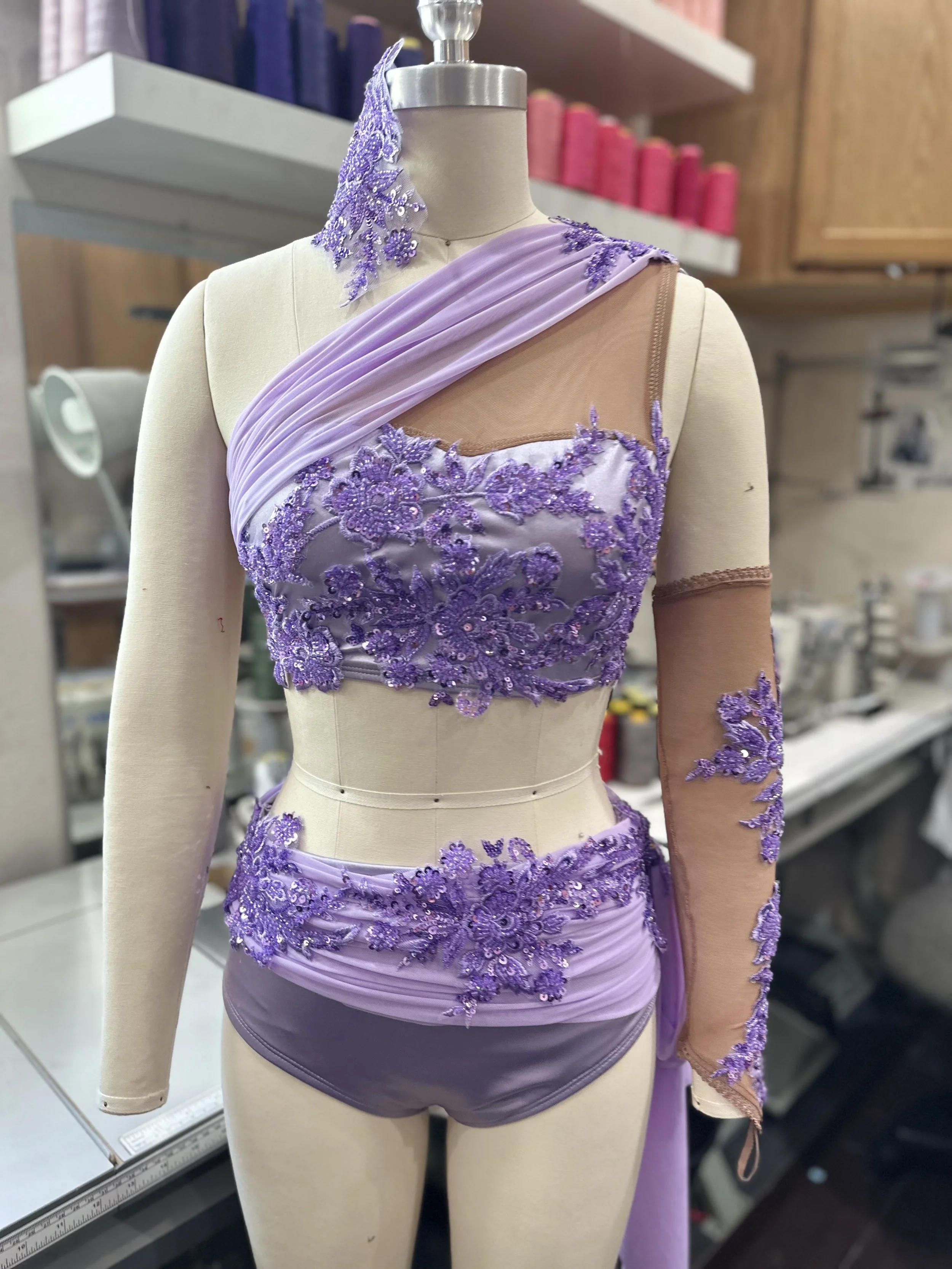A lavender and nude-colored embellished dance costume displayed on a mannequin. The costume features floral embroidery with sequins on the bust, waist, and one sleeve, with a sheer fabric on the other sleeve and diagonal pleats across the chest.