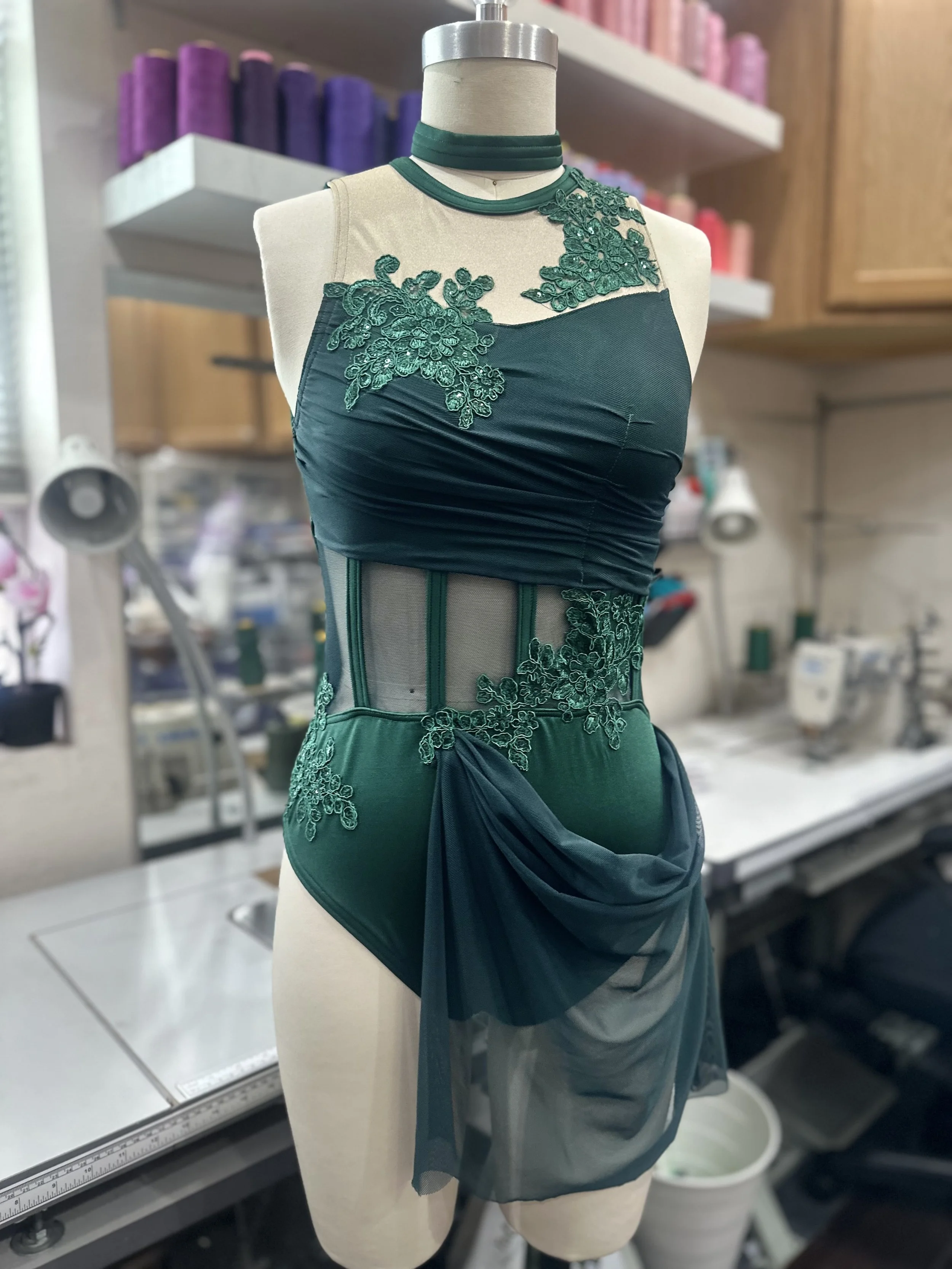 A dress form wearing a dark green, semi-sheer, embroidered dress with floral lace details and draped fabric, in a sewing or tailoring workshop.