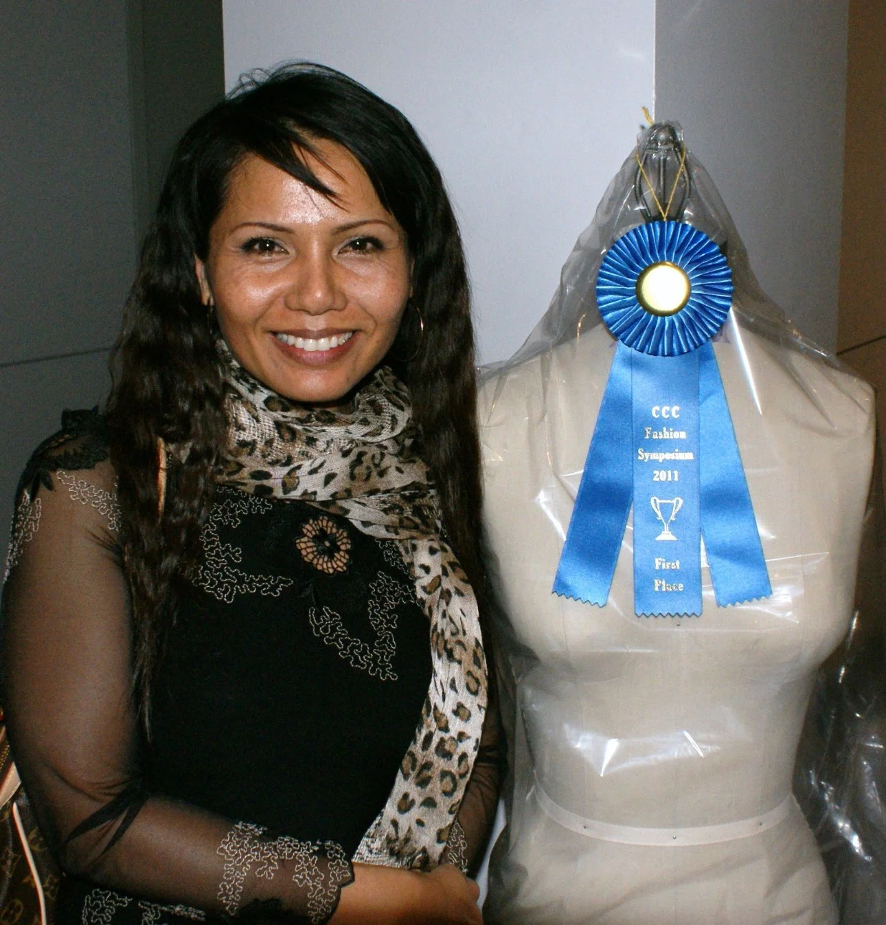 A woman with long dark hair smiling next to a mannequin wrapped in plastic with a blue ribbon that reads 'CCC Fashion Symposium 2011 First Place'.