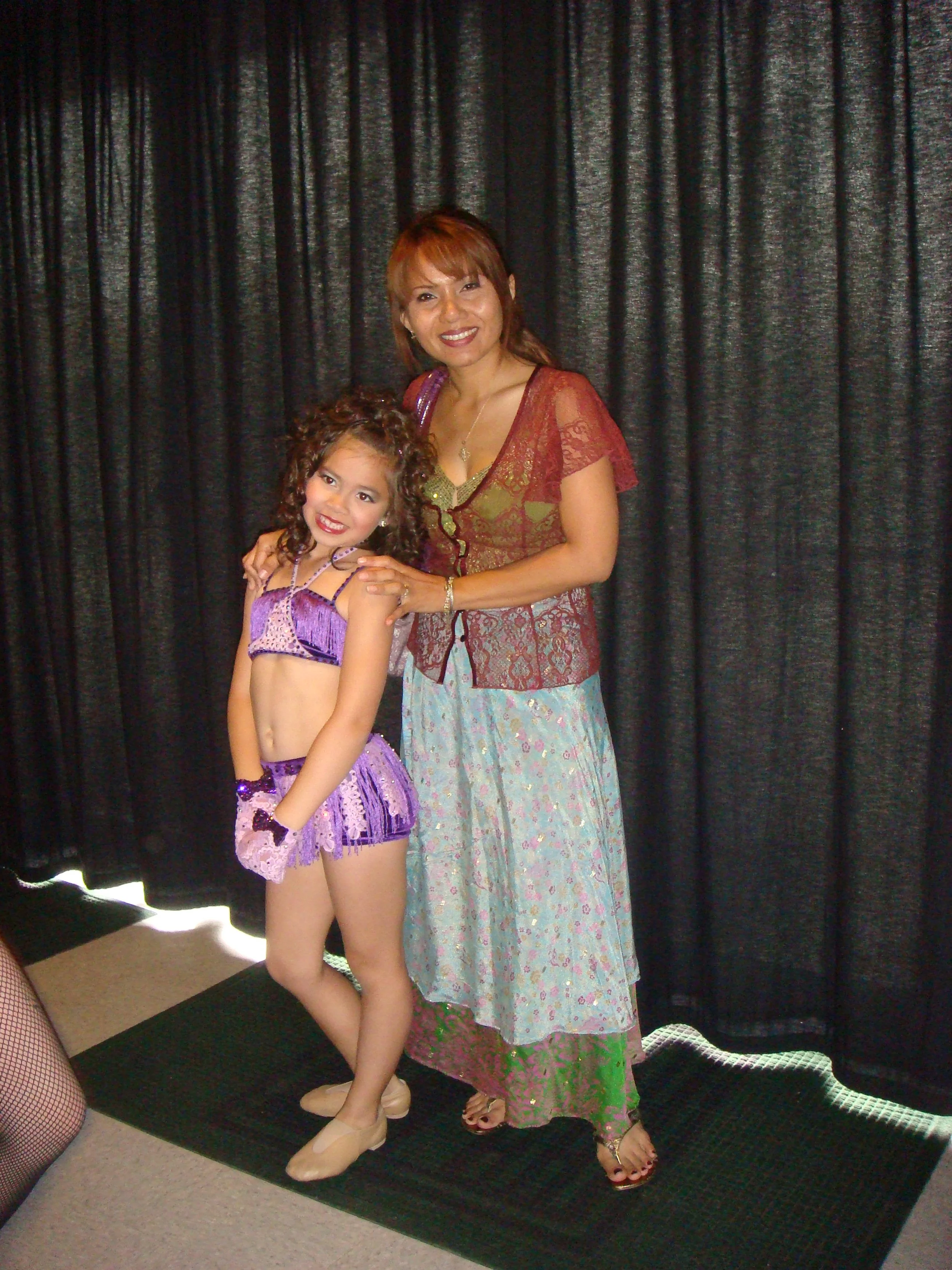 A woman and a young girl standing together in front of black curtains, dressed in dance costumes. The woman is wearing a sheer red lace top with a green shirt underneath, a long patterned skirt, and sandals. The girl is wearing a purple dance costume with fringe and jewelry, and ballet shoes.