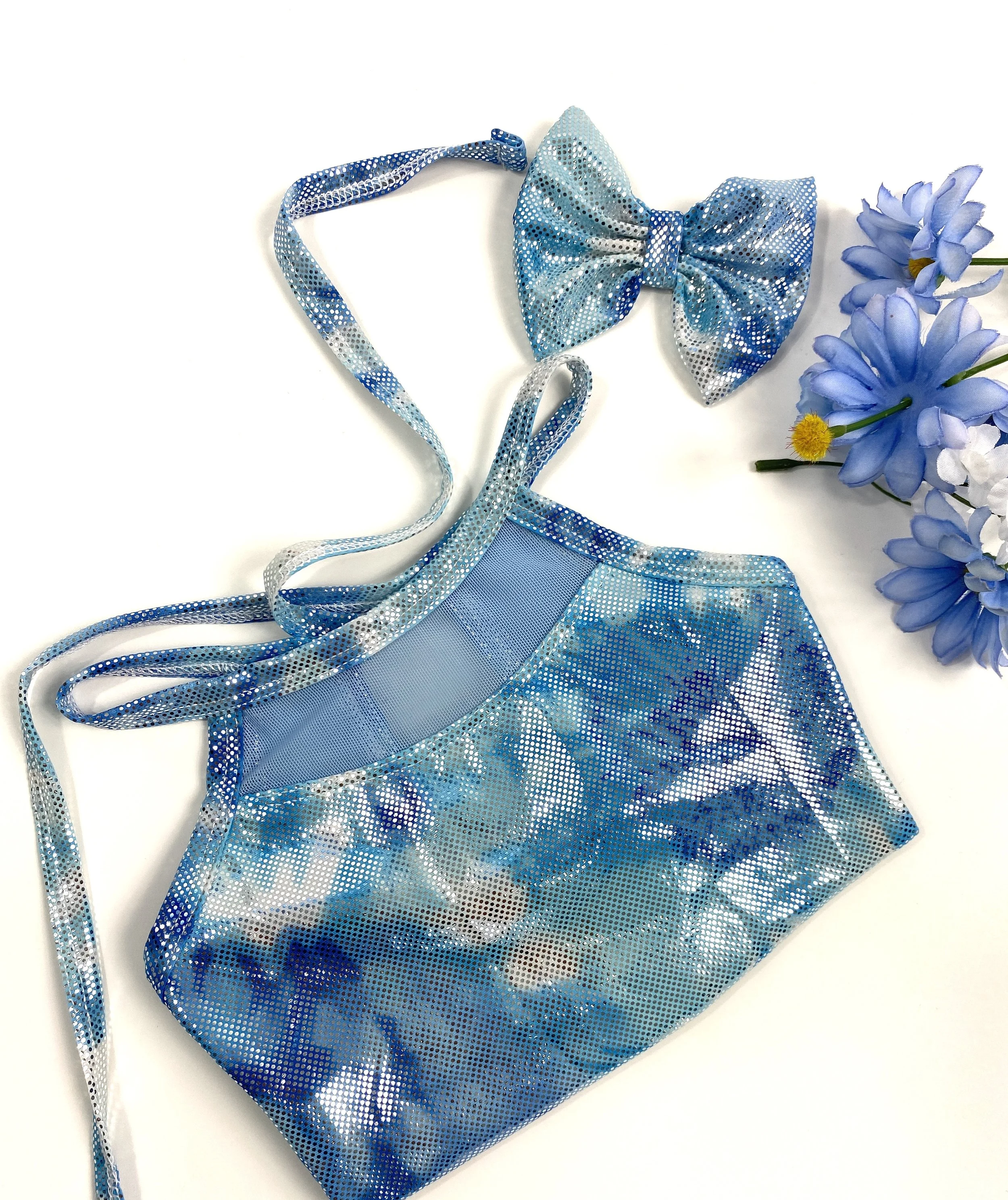 A blue, sparkly mesh handbag with a matching bow and straps, placed next to a bouquet of purple and white flowers on a white background.