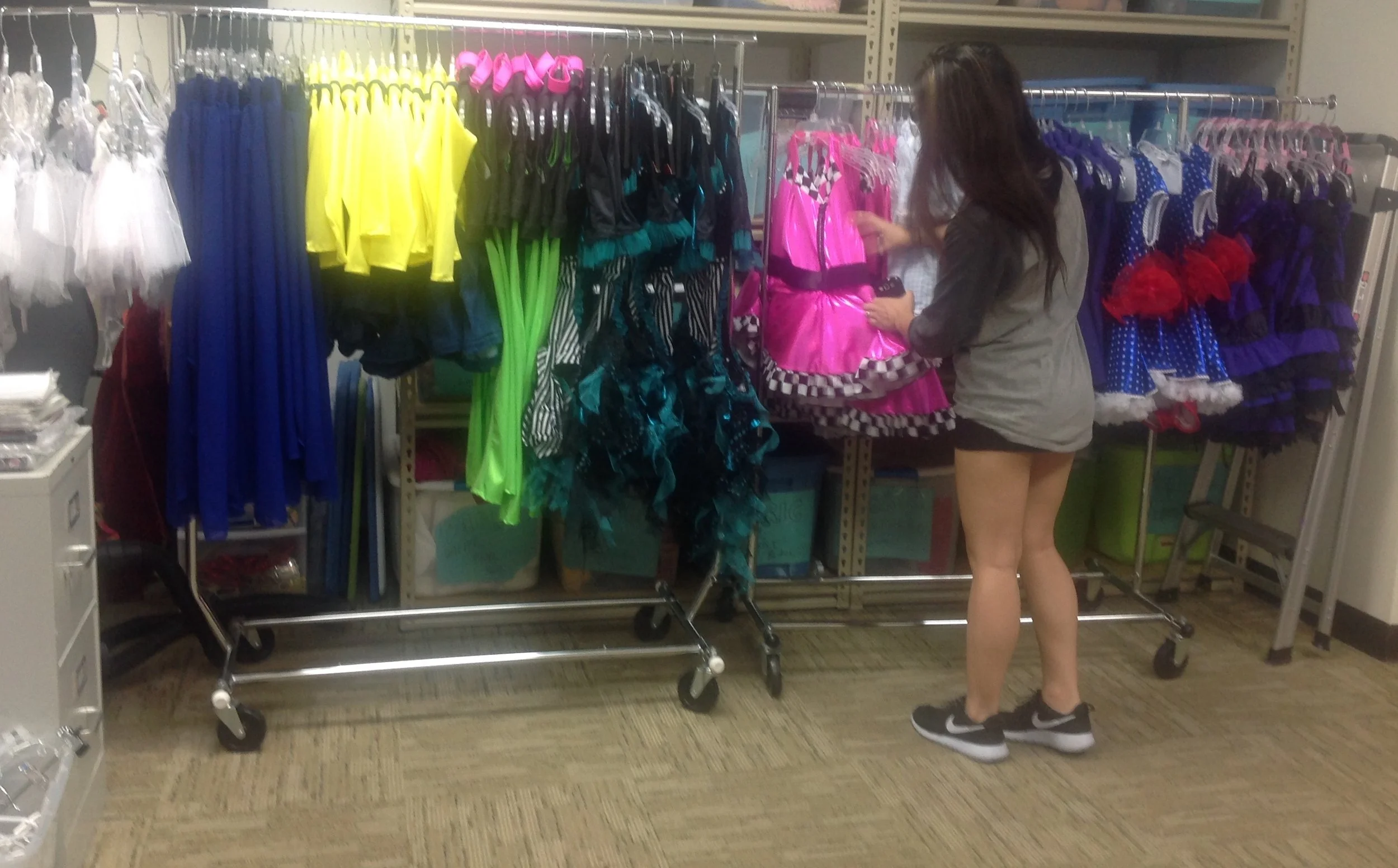 A woman shopping for colorful dance costumes at a store. She is looking at a pink dress with black checkered trim. Racks of ballet and costume dresses in blue, red, purple, yellow, black, and other colors are visible.
