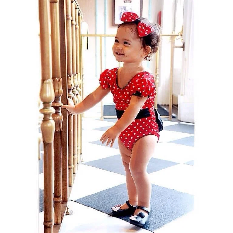 A young girl wearing a red polka dot outfit with a matching headband, black shoes, and a black belt, standing next to a wooden railing indoors. She is smiling and holding onto the railing.
