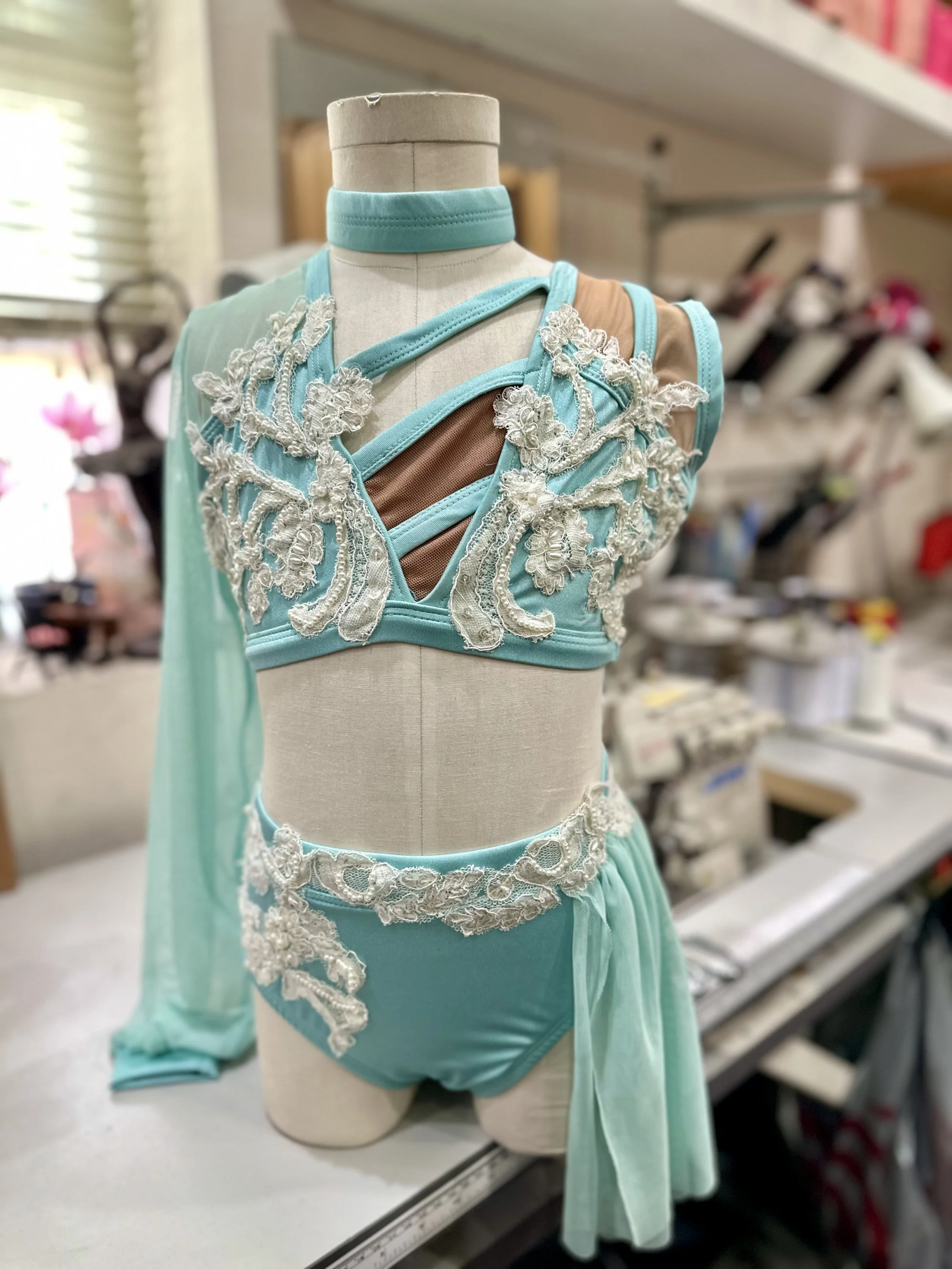 A mannequin dressed in a light blue costume with white lace embroidery and brown accents, displayed on a worktable in a fabric or costume shop.