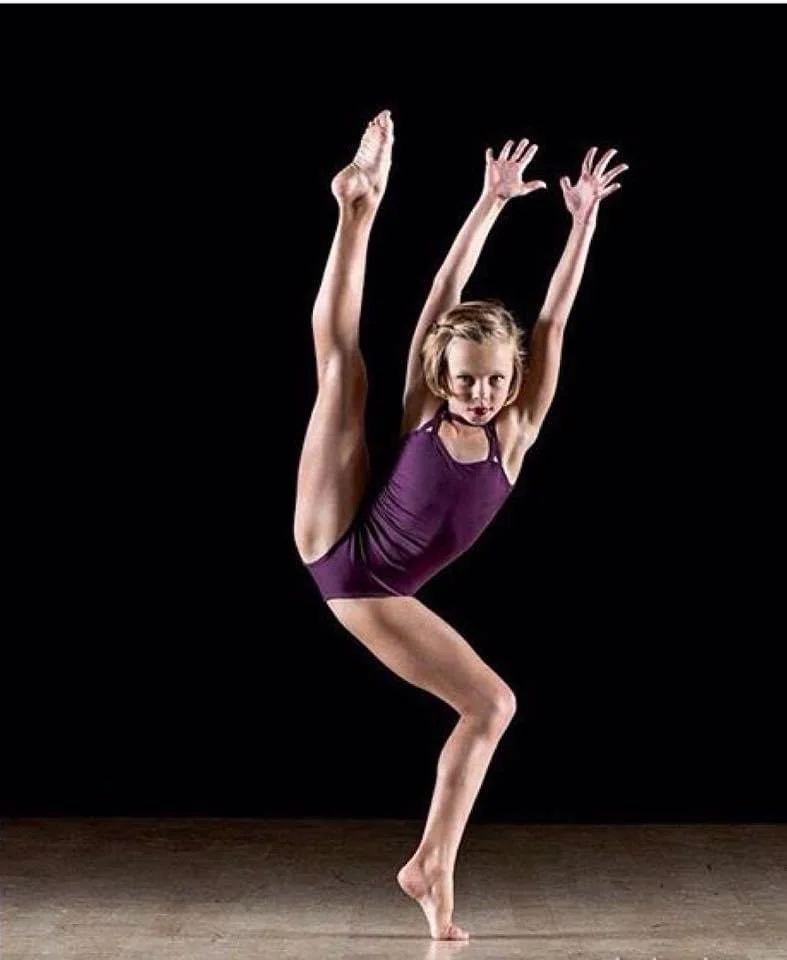 Young girl in purple leotard performing a contorted dance pose against a black background.