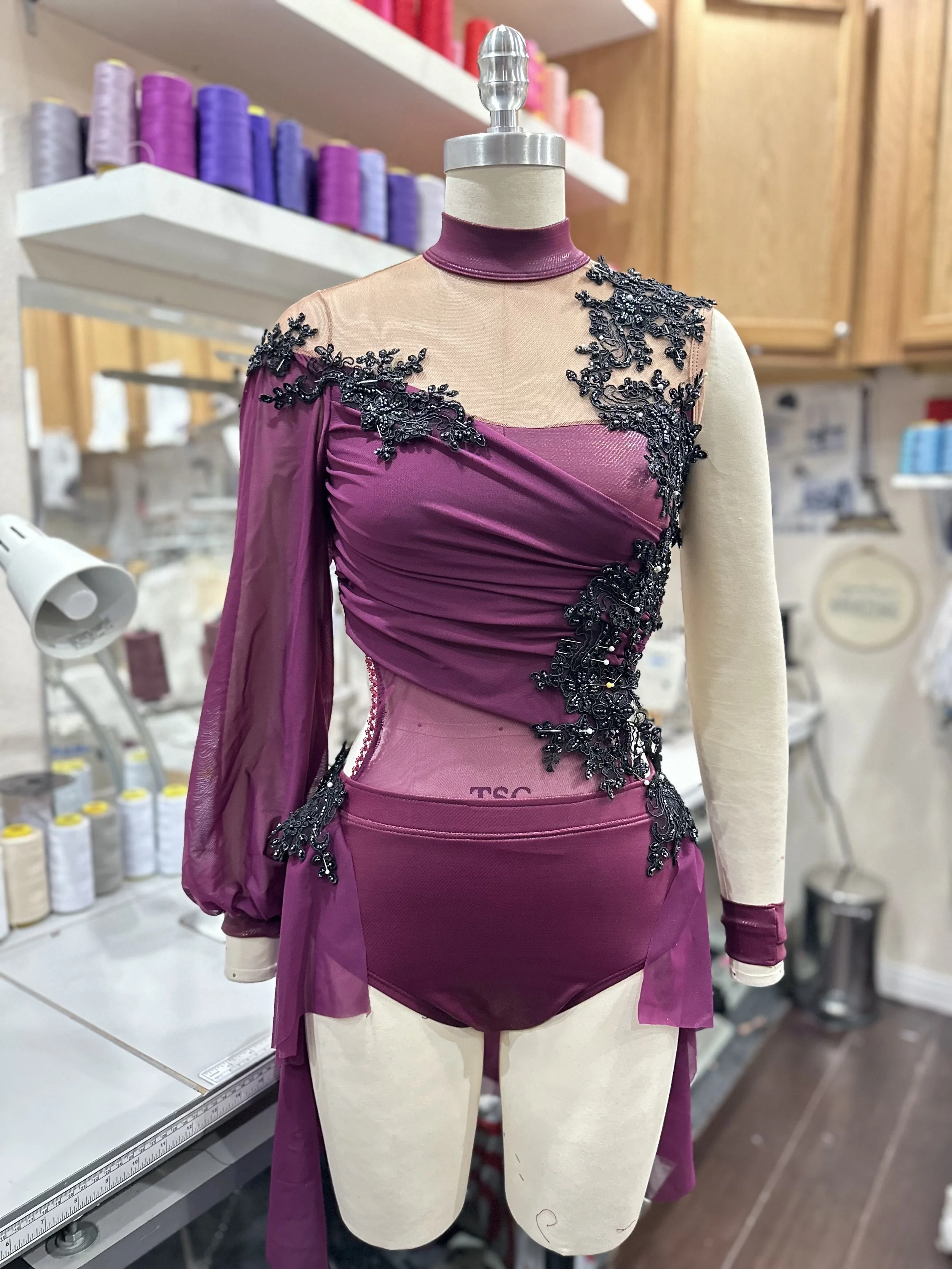 A mannequin displaying a purple and black embellished dress with sheer sleeves, lace details, and a high neckline, placed in a textile or clothing workshop.