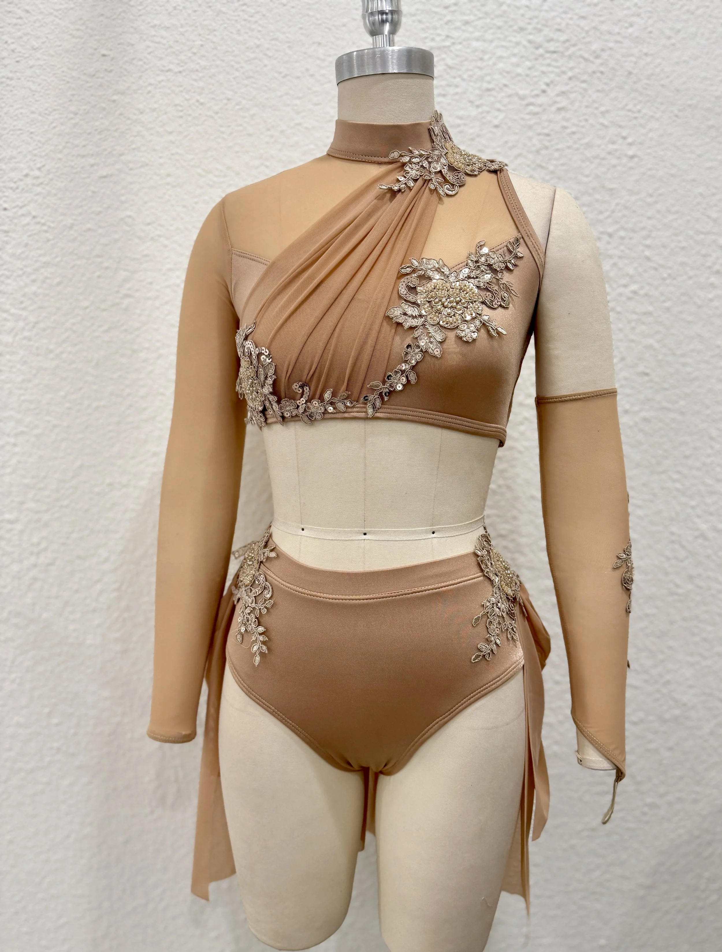 A mannequin dressed in a beige and tan embroidered dress with embellishments, standing against a plain wall.