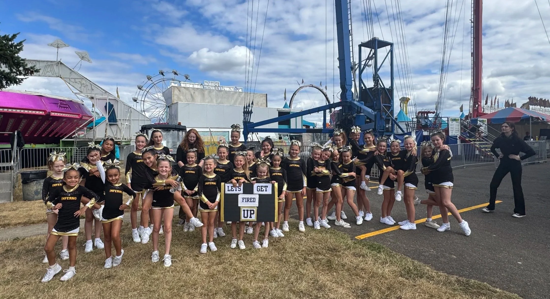 Group of young cheerleaders at the Clark County Fair in matching black and gold cheerleading uniforms outside at a fair, standing in front of amusement rides and holding a sign that reads 'Let's Get Fired Up. camas, washougal, Vancouver ridefield