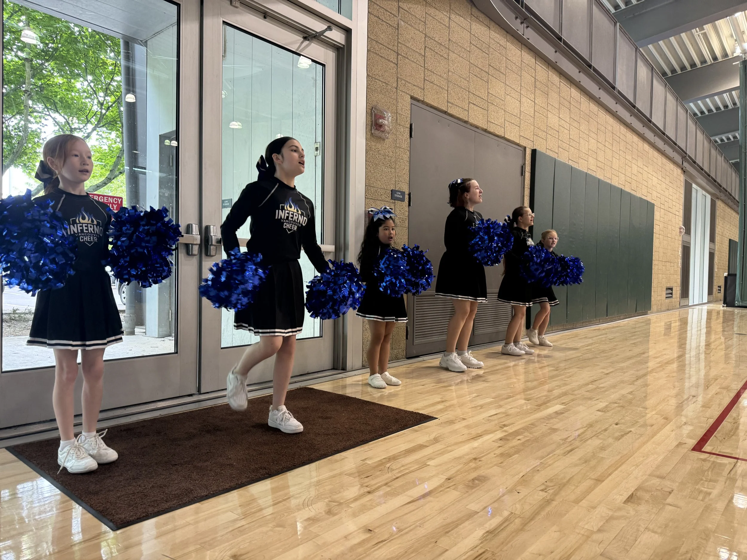 A group of young cheerleaders standing inside a gymnasium, holding blue pom-poms. They are dressed in black and white cheerleading uniforms, some with hair tied back, and are standing near a glass door and a wall, prepared for a cheerleading routine.