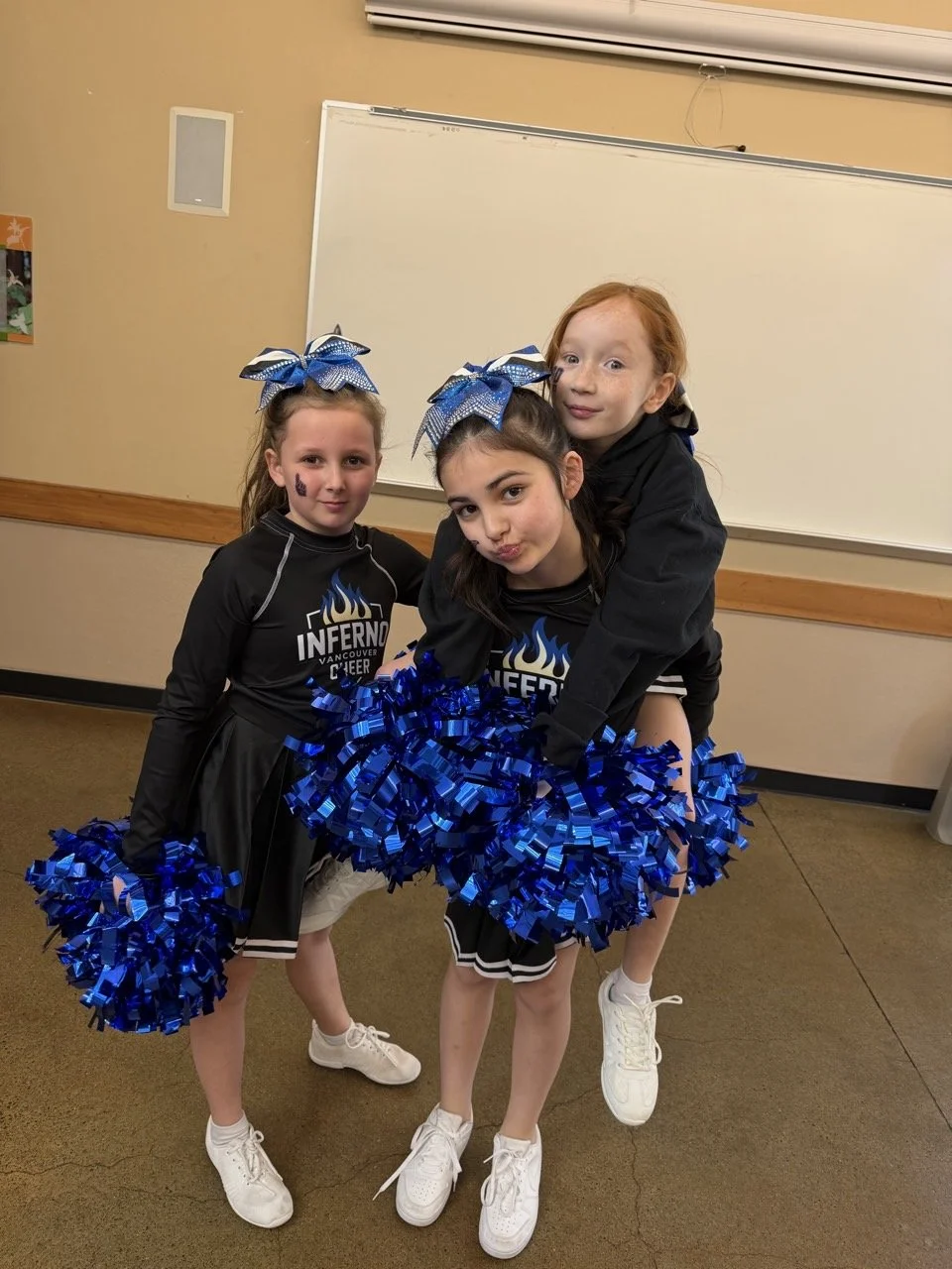 Three young cheerleaders with blue pom-poms posing indoors. They are wearing black cheerleading uniforms with blue bows in their hair, and one girl is riding on another's back.