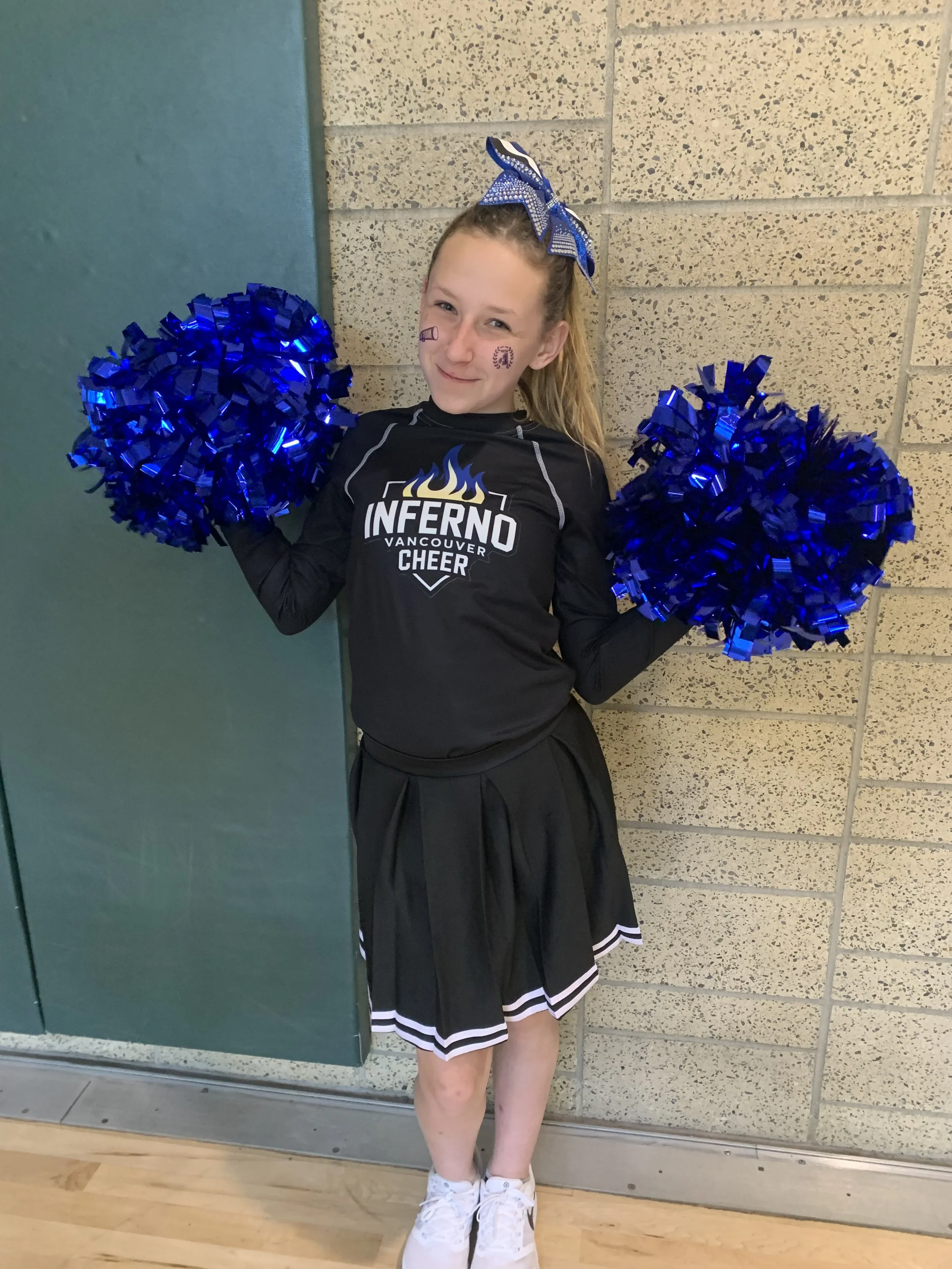 Young girl dressed as a cheerleader with blue pom-poms, a blue bow in her hair, and face stickers, standing against a brick wall indoors.